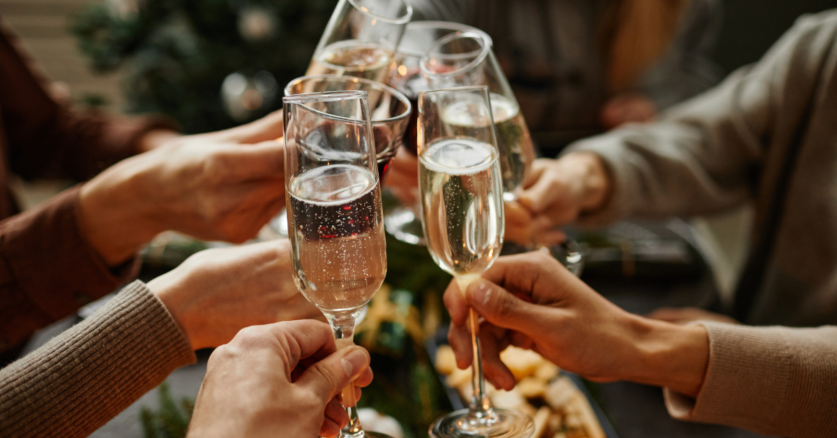 Several hands raising champagne flutes in a celebratory toast during a holiday gathering, with twinkling lights and seasonal décor softly blurred in the background to create a warm festive atmosphere.