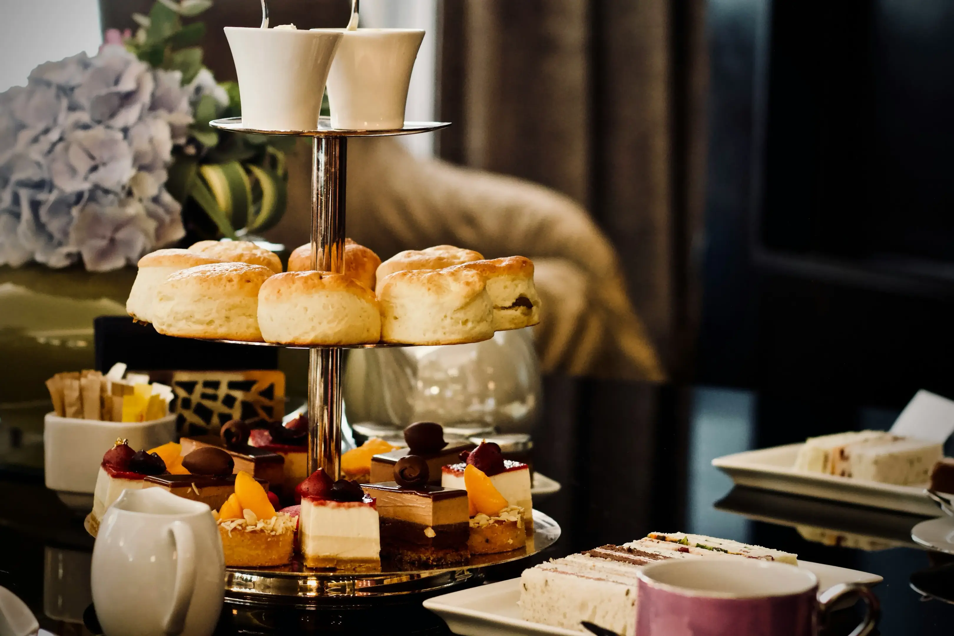 A three-tiered serving stand is filled with afternoon tea treats, featuring fresh scones on the middle tier and a colorful assortment of pastries, tarts, and sliced cakes on the bottom. In the foreground, plates of finger sandwiches, a tea pitcher, and a teacup rest on a dark, reflective table alongside a floral arrangement.