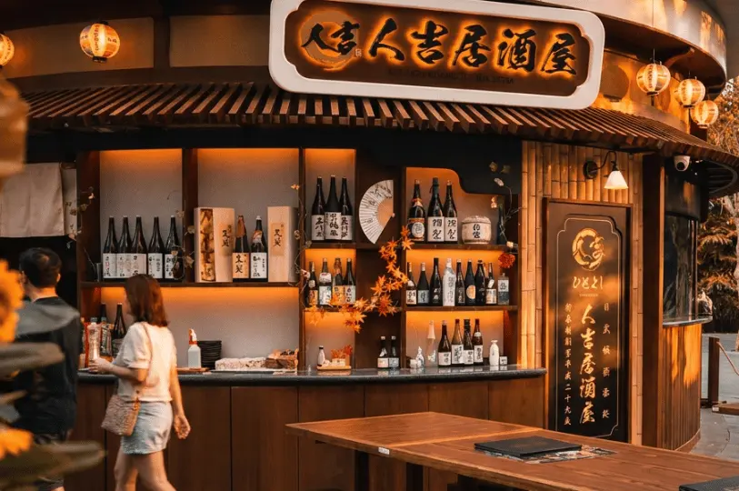 The warmly lit exterior of an izakaya showcasing traditional wooden architecture, a prominent signboard, and an outdoor display shelf heavily stocked with various premium sake bottles.