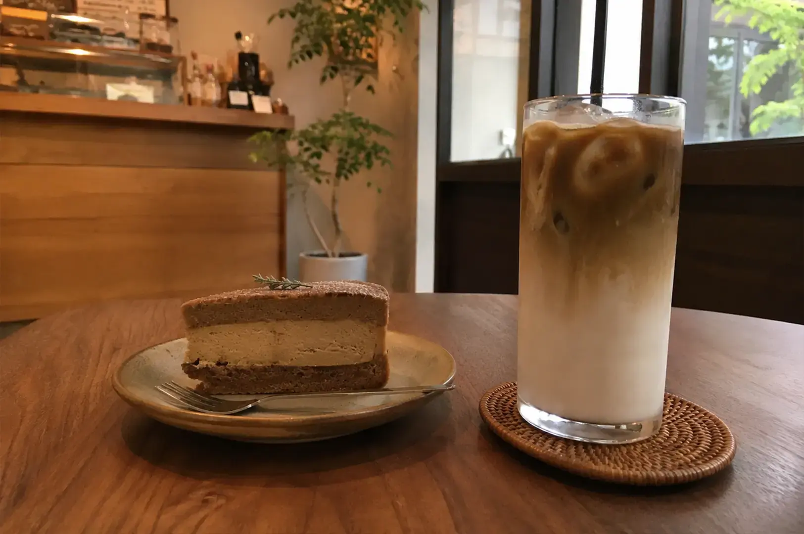 A slice of layered cake topped with a small twig garnish sits on a ceramic plate next to a tall glass of iced layered drink, all resting on a dark wooden table in a cozy cafe setting.