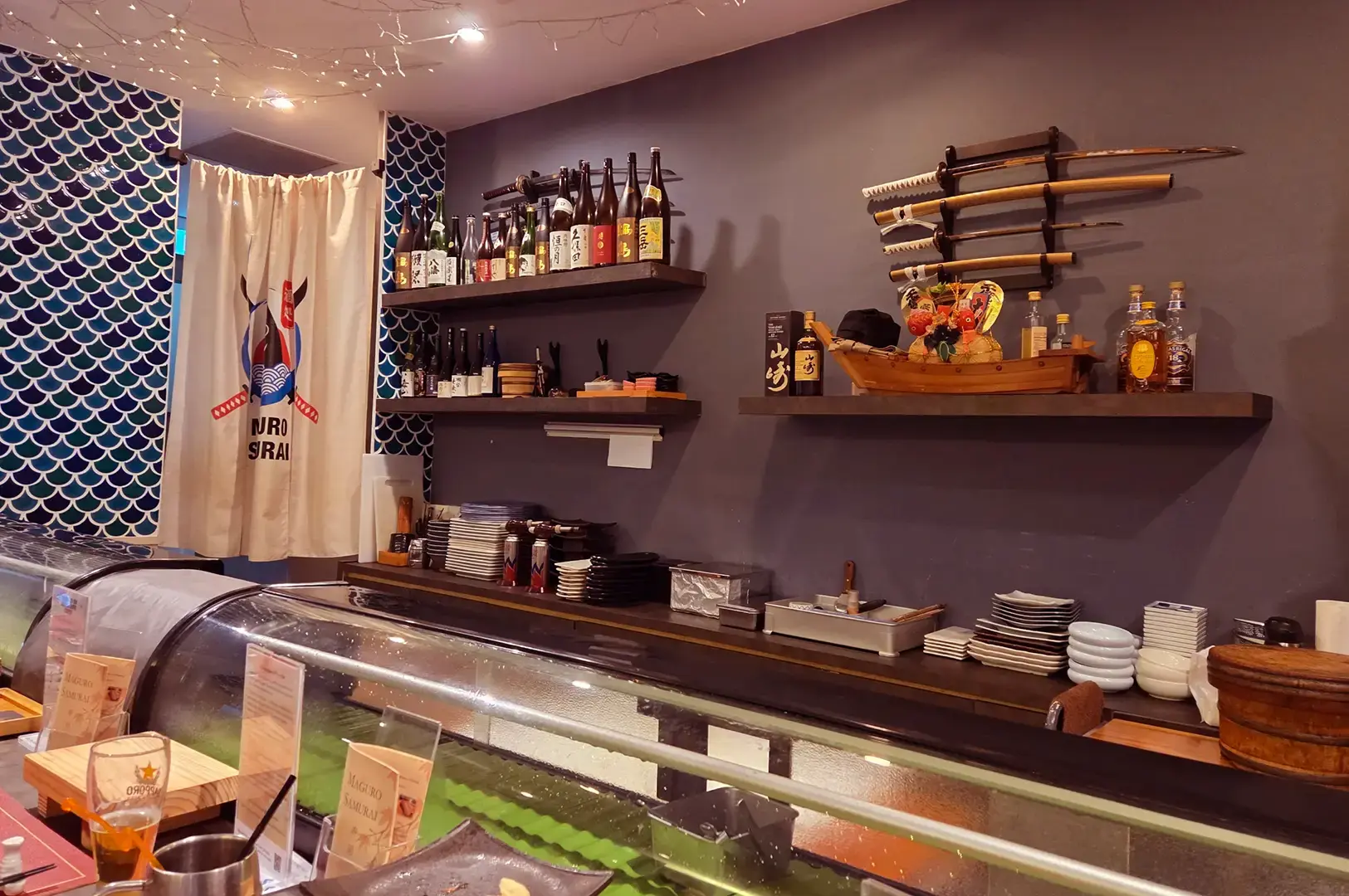 Maguro Samurai sushi bar with a refrigerated display counter, shelves of sake and spirits, stacked plates, and Japanese decorations.