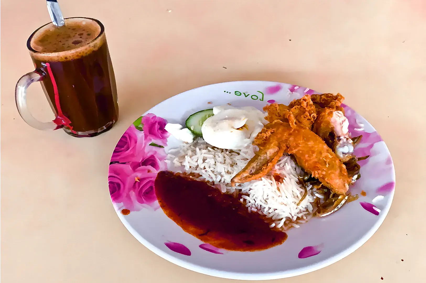 Plate of nasi lemak with fried chicken, rice, sambal, sliced cucumber, and boiled egg on a floral plate. A glass of tea is beside it.