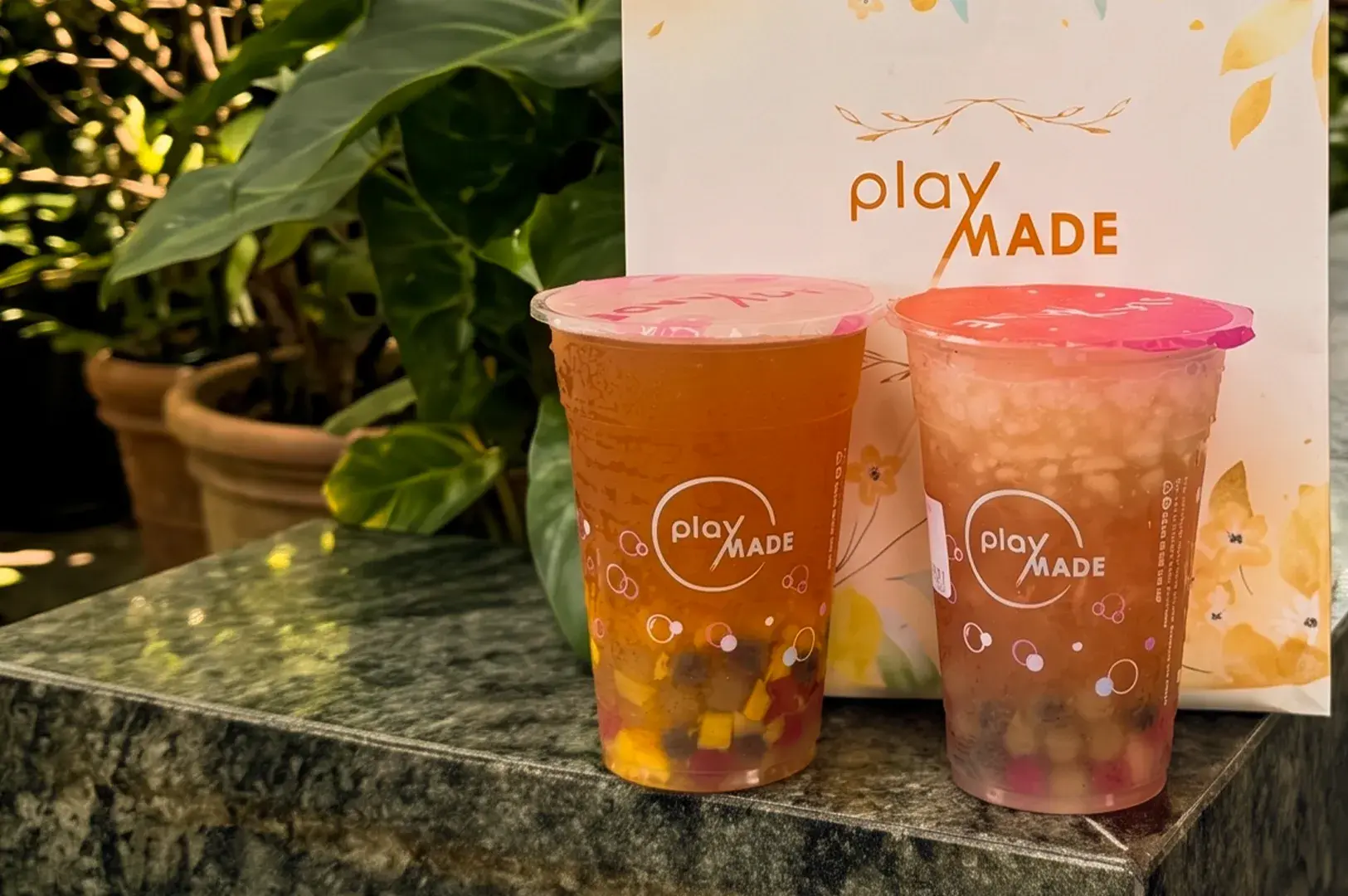 Two branded PlayMade bubble tea cups with colorful toppings sitting on a speckled countertop next to a matching gift bag.