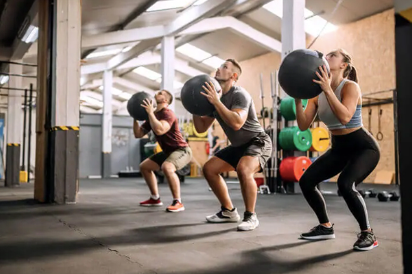 Group fitness class doing squat-to-throw medicine ball reps, training physical fitness components health related like strength and cardiovascular endurance.