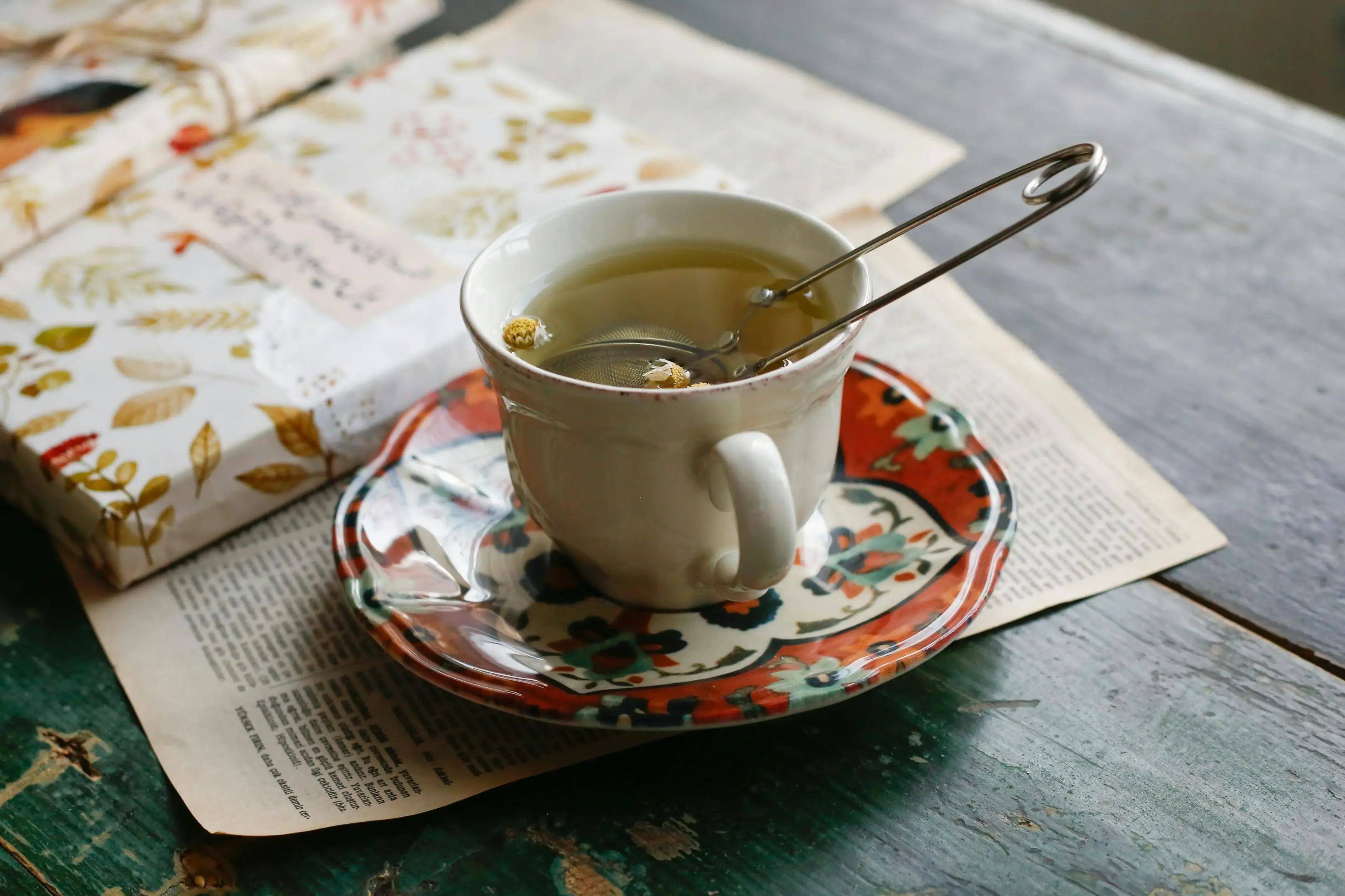 A teacup filled with chamomile tea and a mesh infuser rests on a colorful, patterned saucer over an open book. In the background, a wrapped book with a floral pattern sits on a weathered green wooden table.