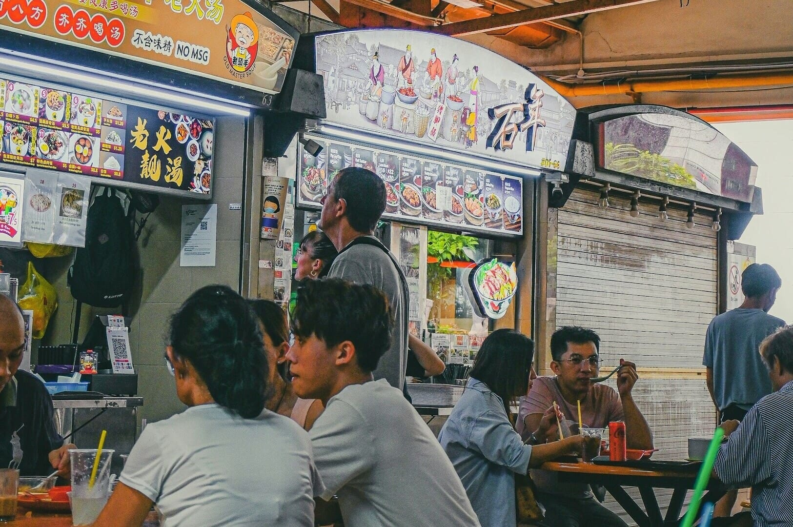 Diners seated at tables, savoring dishes from a bustling Singapore hawker food stand.
