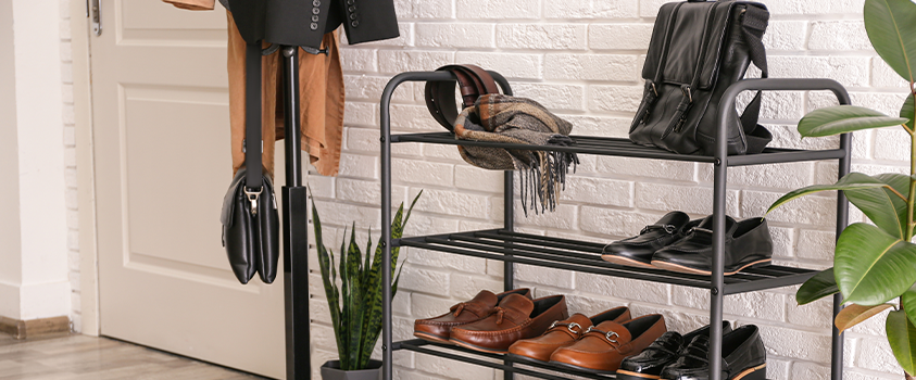 An entryway with light wood floors and white brick walls. Right of centre is a black metal shoe rack holding a variety of shoes, a black leather handbag, a scarf and a belt. To the left of the rack is a pot plant and a black coat rack.