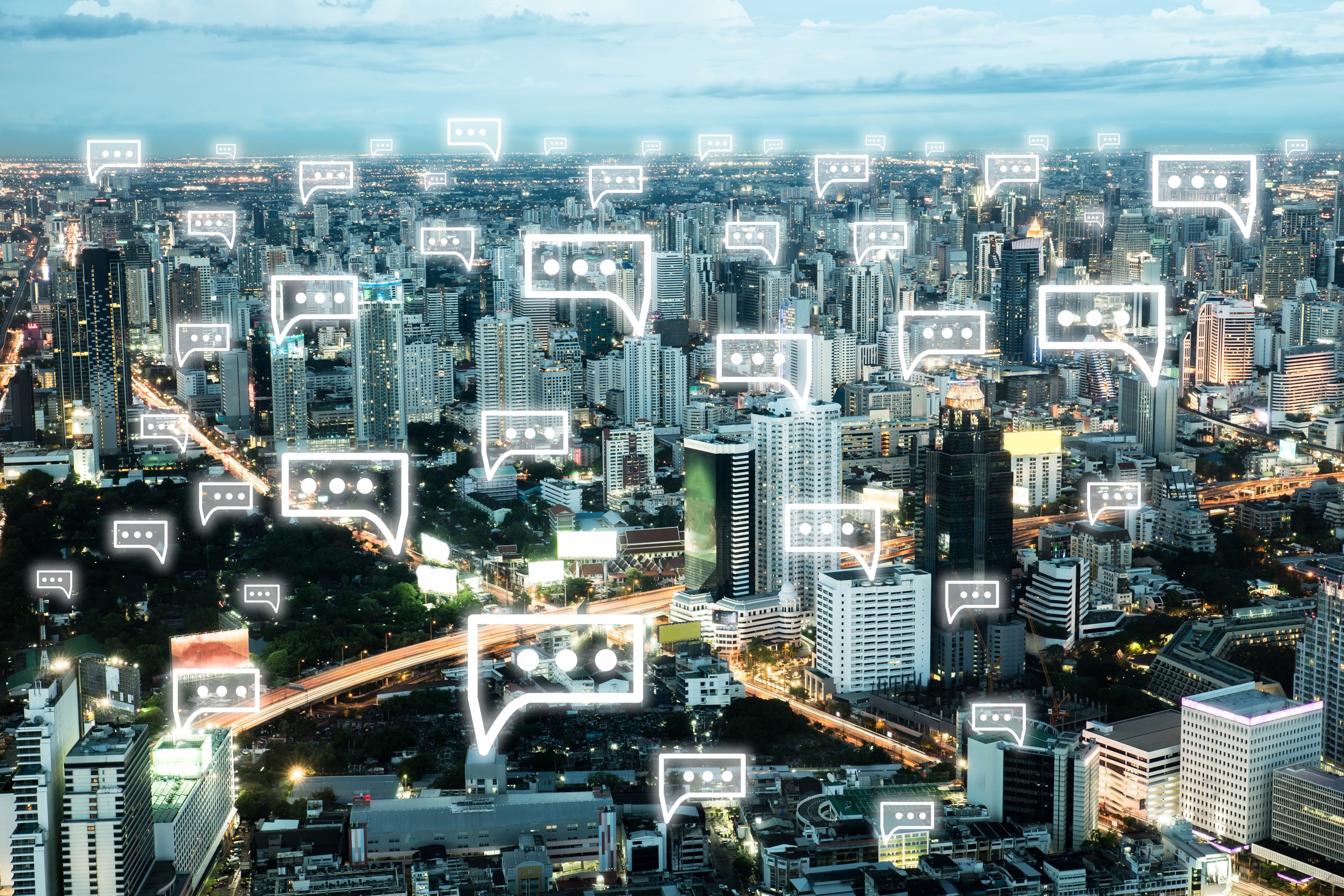 An aerial view of a modern cityscape at dusk, with dozens of glowing speech bubble icons floating above buildings. The visual illustrates how UCaaS solutions and cloud-based communications enable continuous, real-time unified communications in densely connected urban environments.
