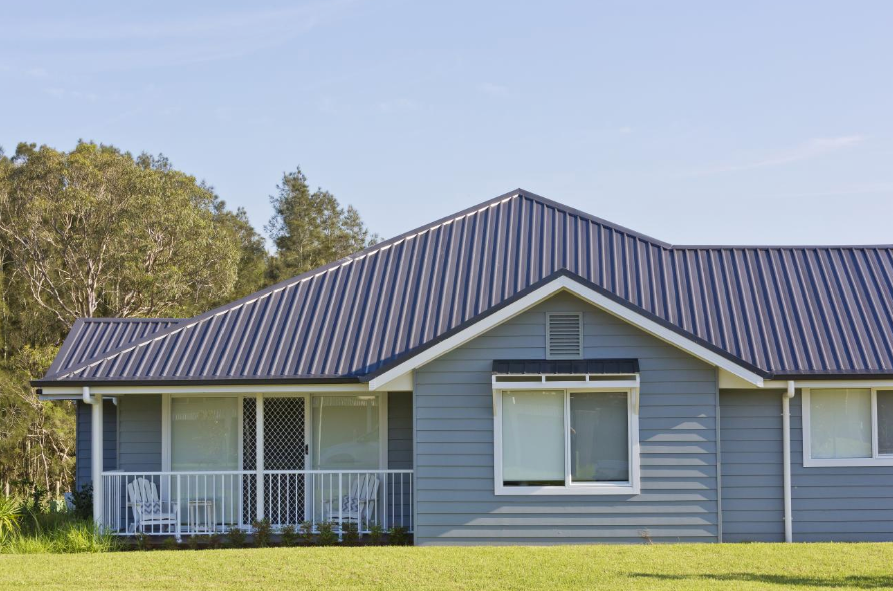 A modern house with a light blue exterior and a roof featuring dark blue Trimdek roofing sheets. The house has white-framed windows and a small porch with white railings, set against a backdrop of green lawn and trees