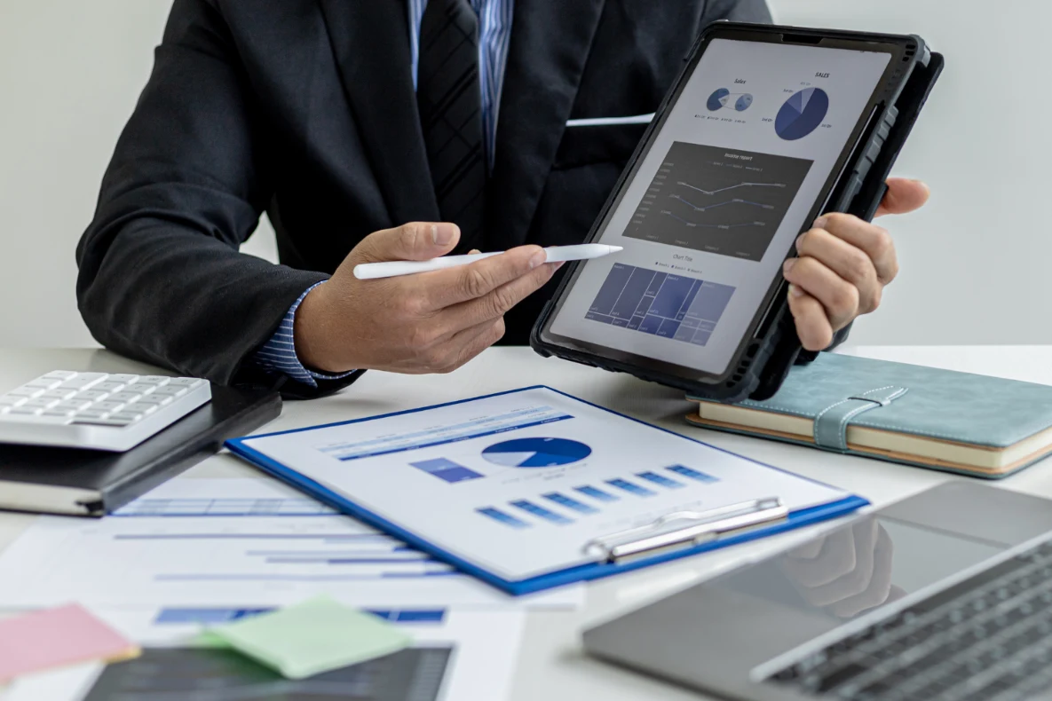A person in a suit points to a tablet displaying charts and graphs, with a clipboard, calculator, and laptop on the desk.