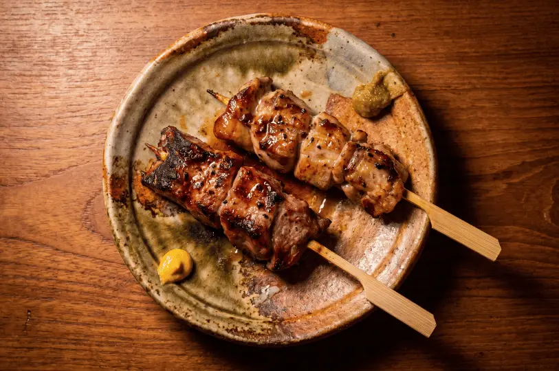 A close-up of two grilled meat skewers with a dark glaze, resting on a rustic, earthy ceramic plate alongside small dabs of yellow mustard and green condiment, set on a wooden table.