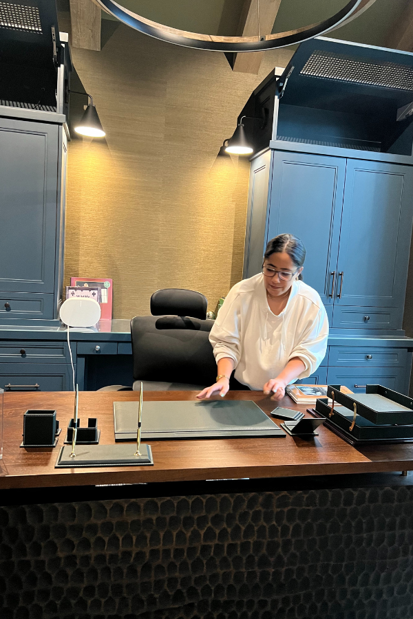 A designer arranges materials on a large wooden desk in a dark, sophisticated office with blue built-ins and warm lighting.