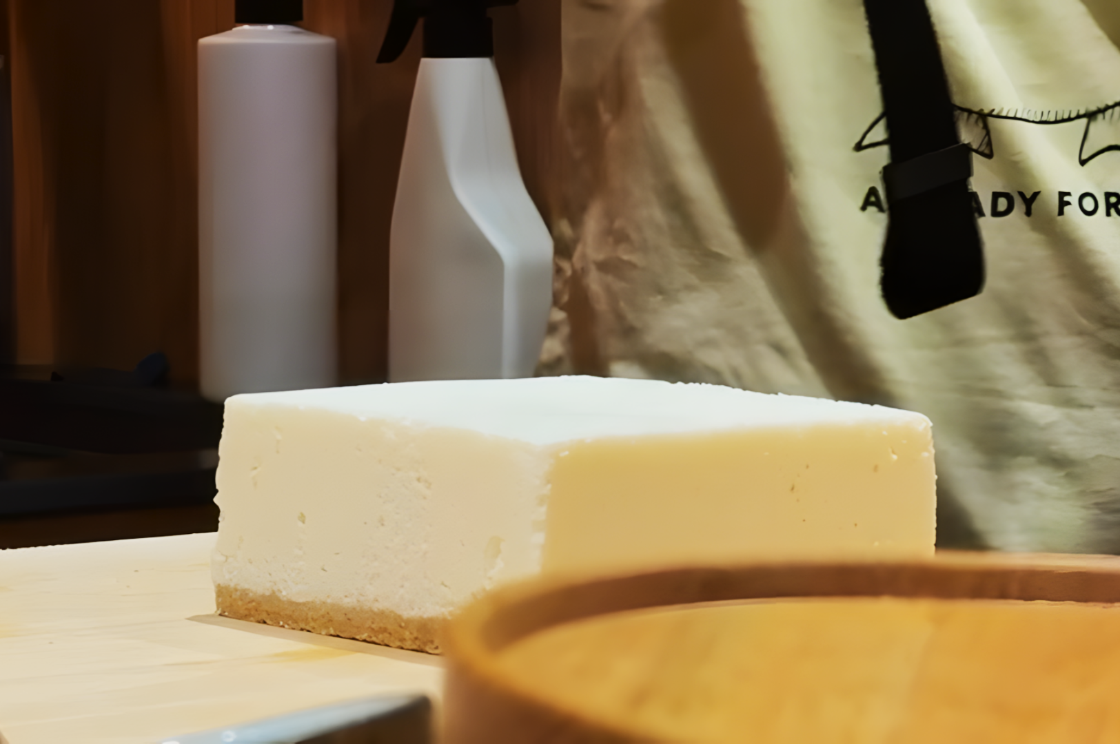 A block of white tofu on a wooden cutting board in a kitchen setting. A spray bottle is in the background, with part of a person visible nearby.