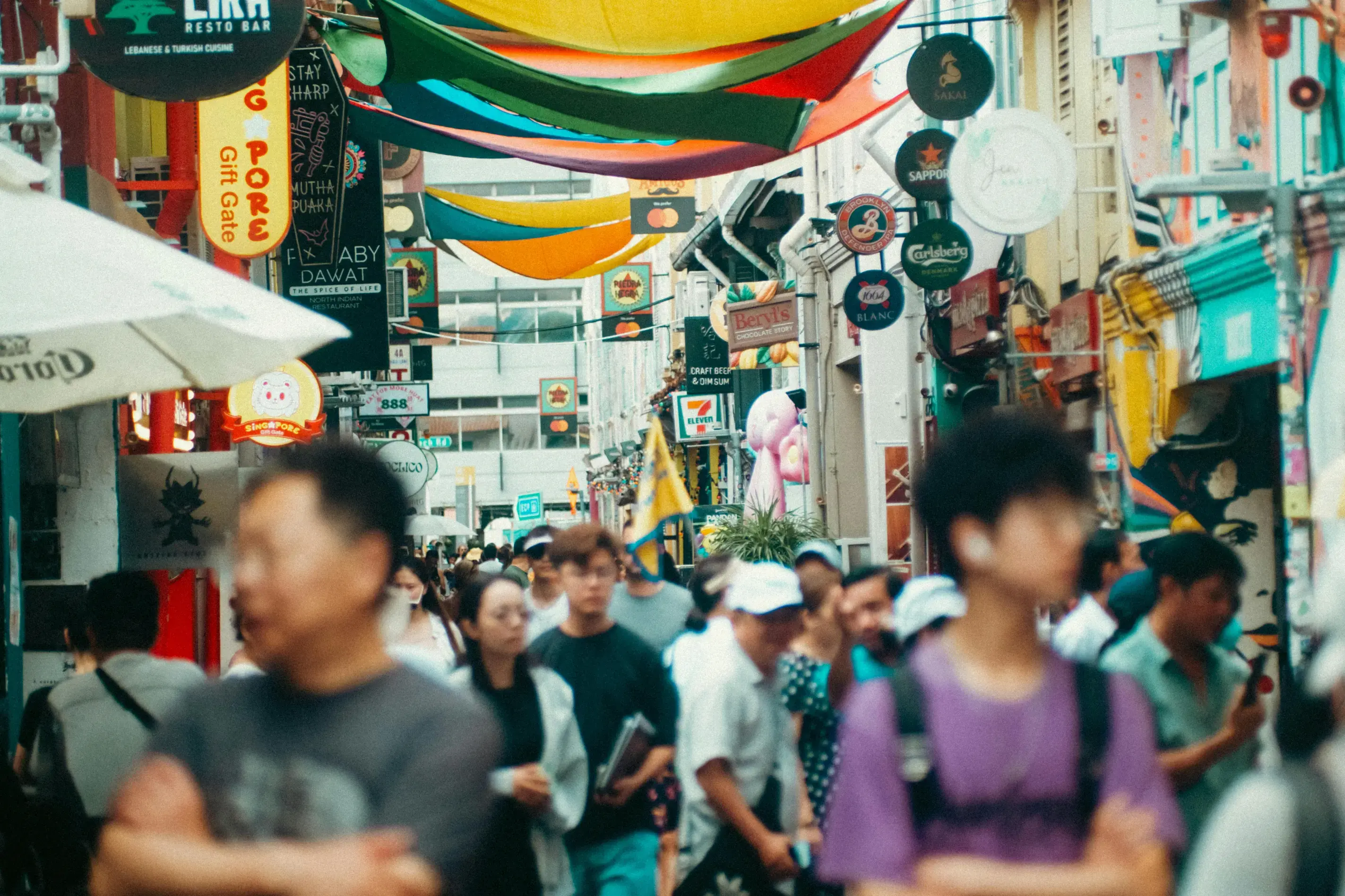 A vibrant and bustling narrow street is filled with people walking beneath a canopy of colorful, wave-like banners. Numerous eclectic shop signs, including those for restaurants and convenience stores, line the sides of the street, creating a dense and lively urban atmosphere.