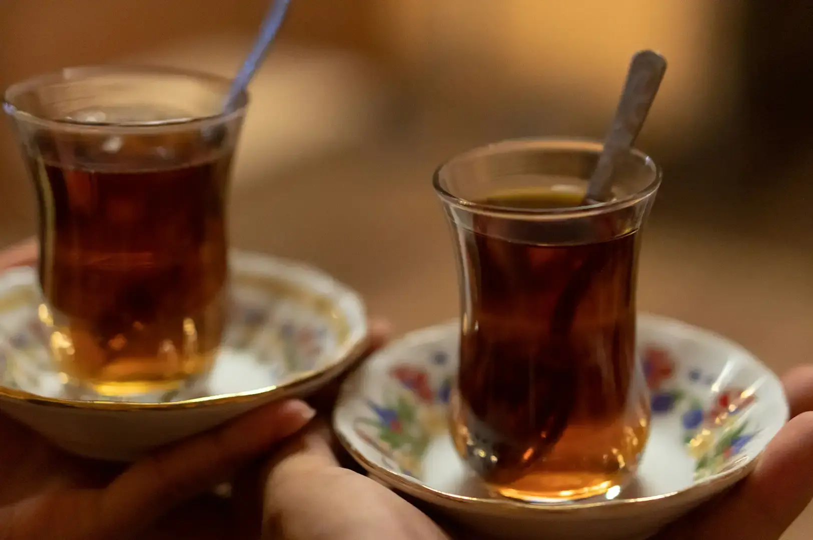 Two hands delicately hold up two tulip-shaped glasses filled with hot, dark amber Turkish tea, each resting on its own floral-patterned ceramic saucer with a small spoon inside, presented against a soft, warm-toned, blurred background.