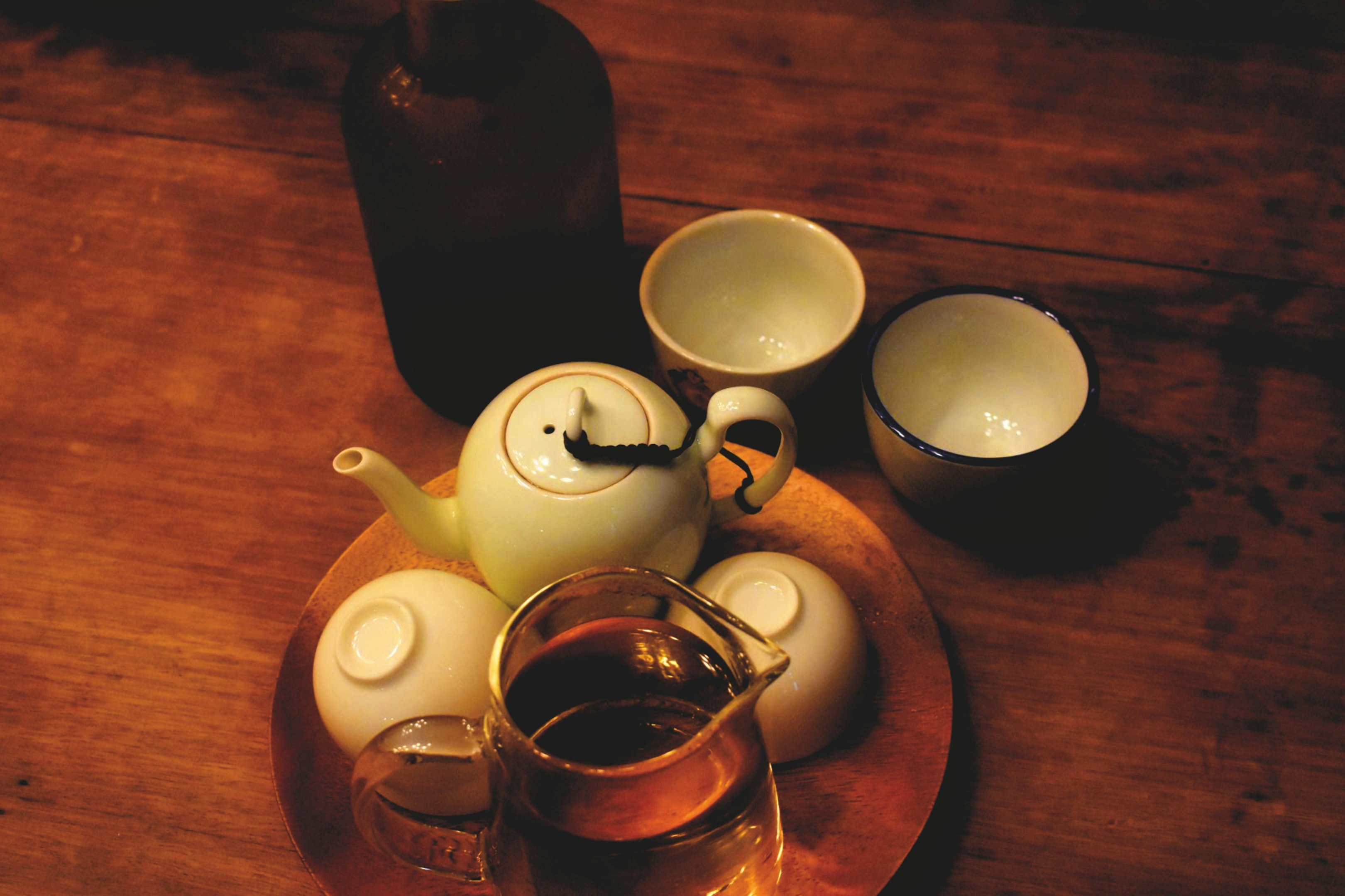 A warm, dimly lit scene shows a traditional tea set including a small ceramic teapot, several cups, and a glass pitcher of tea arranged on a wooden table. A dark bottle stands in the background, adding to the cozy and rustic atmosphere of the setting.