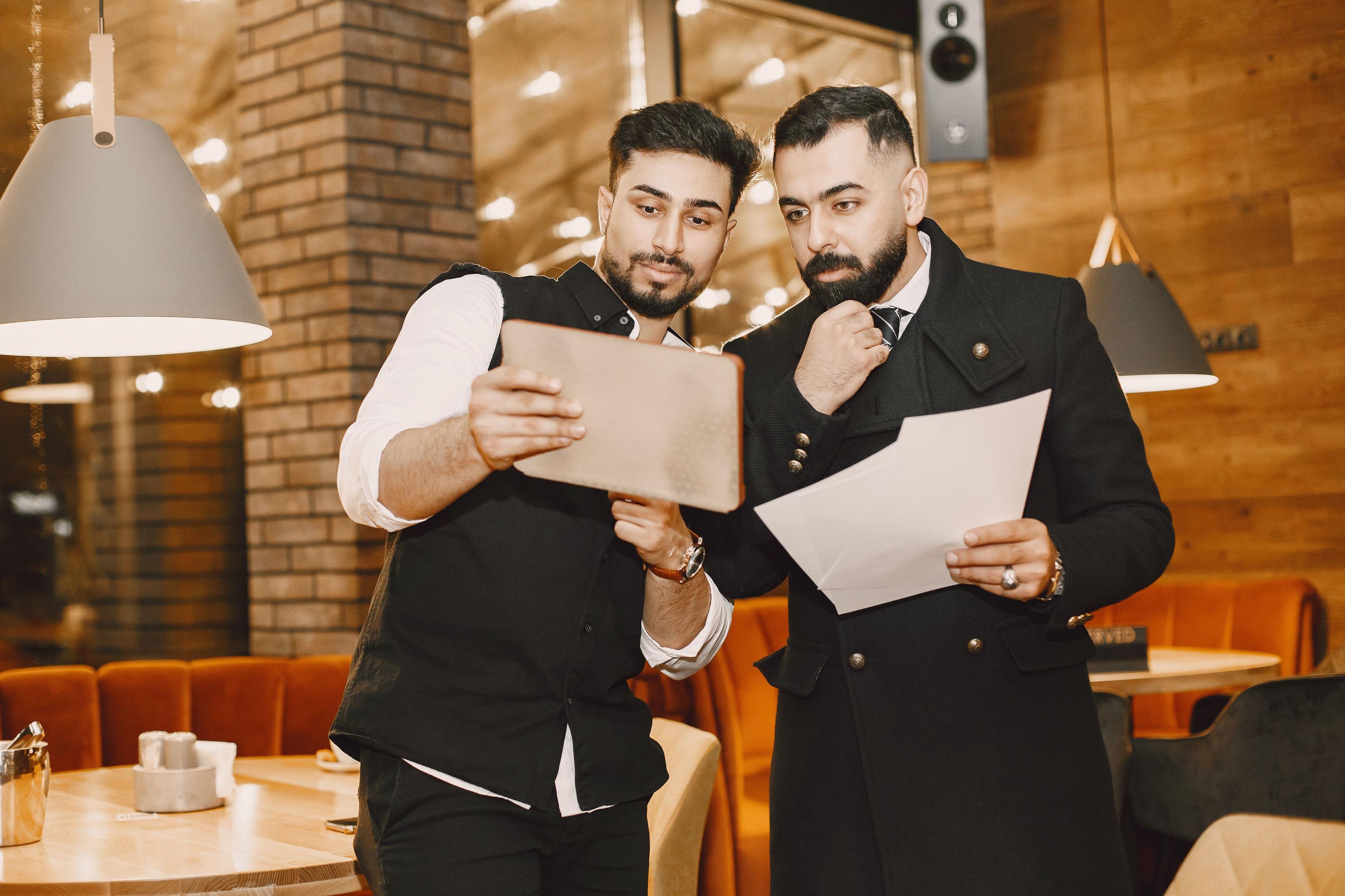 A restaurant team is gathered near the kitchen pass, reviewing customer orders on a tablet, utilizing AI tools to enhance operational efficiency and customer satisfaction in the restaurant setting. This collaborative effort aims to streamline the ordering process and improve the dining experience by leveraging data-driven insights.