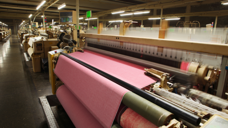 A large industrial loom weaving striped pink and white fabric in a factory setting.