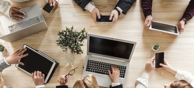 Ujęcie z góry pokazuje dłonie ludzi pracujących na laptopach, telefonach i tabletach.