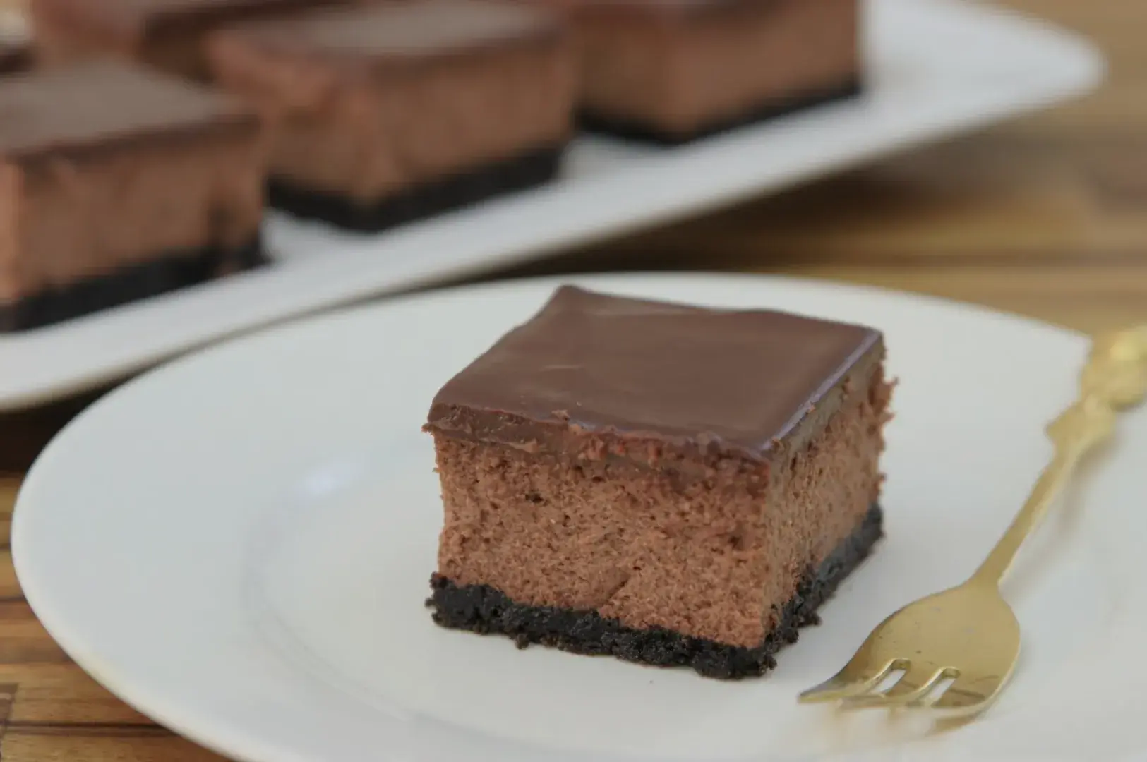 A slice of chocolate cheesecake with a chocolate ganache topping on an Oreo crust, served on a white plate with a gold fork.