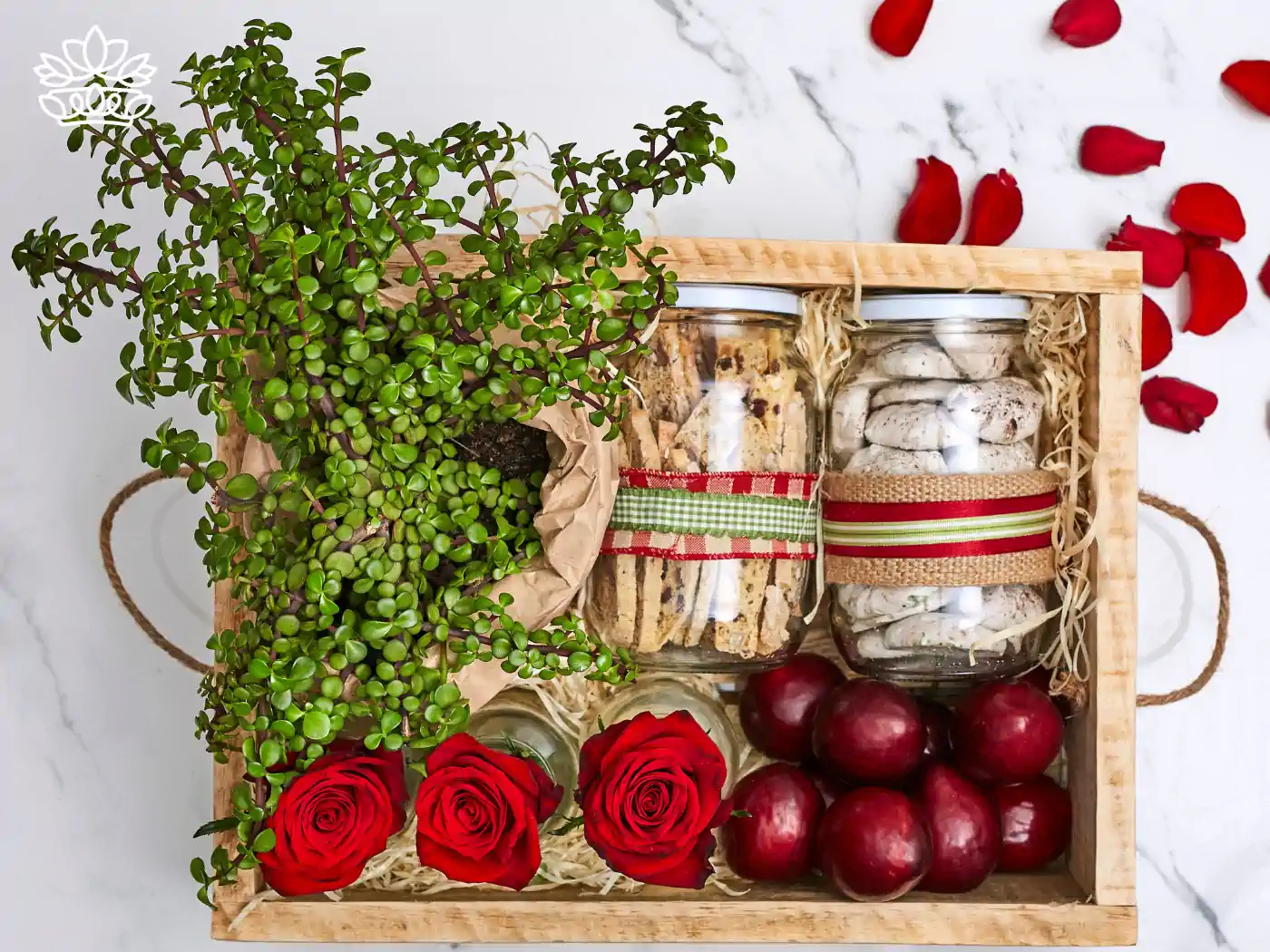 A beautifully arranged gift box with a vibrant potted plant, fresh red roses, jars of homemade cookies and meringues, and lush red apples, set against a marble background with scattered rose petals. Guest House and Hotel Gift Boxes Delivered with Heart. Fabulous Flowers and Gifts.