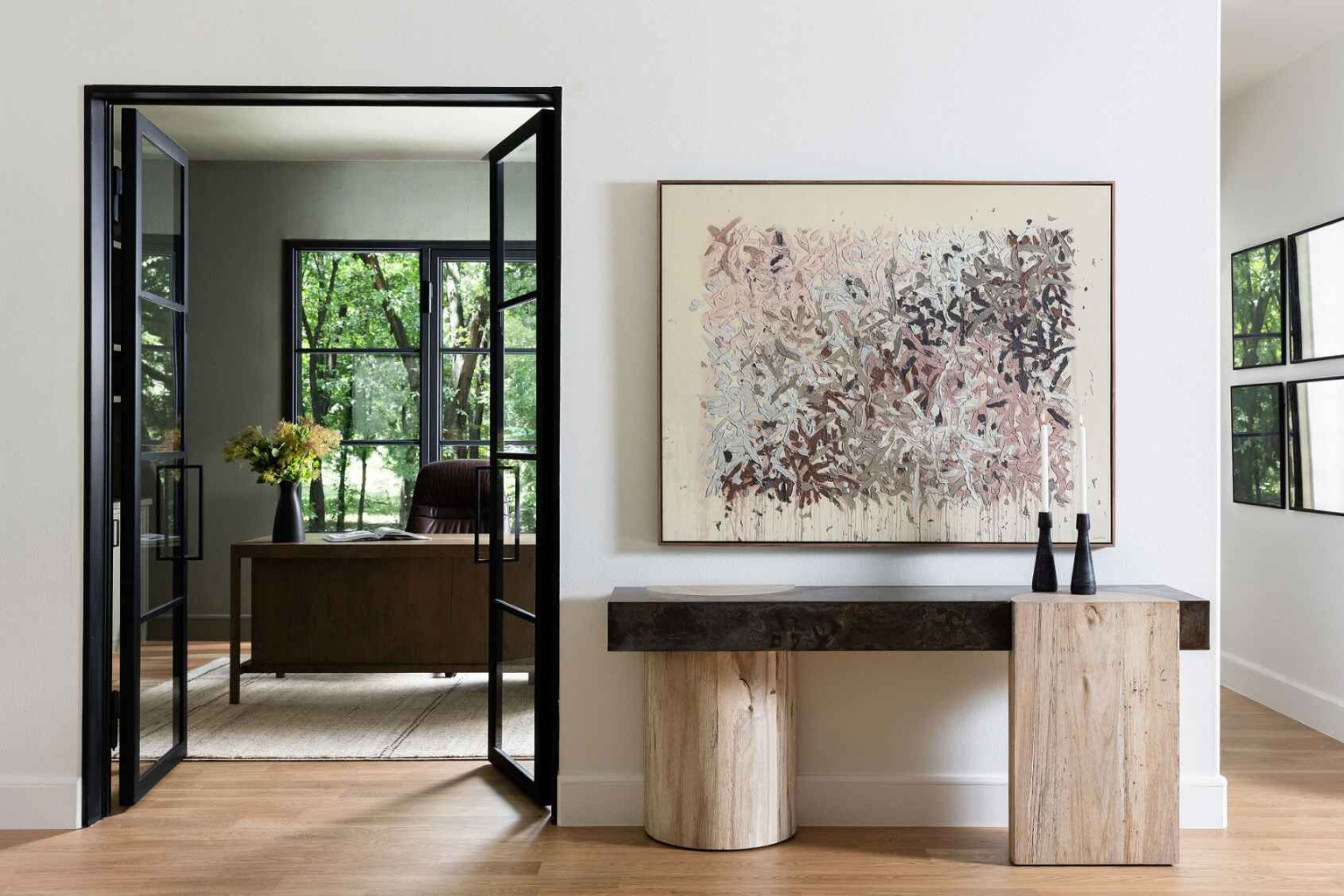 Minimalist hallway with a dramatic abstract painting, sculptural console table, and black French doors leading into a home office filled with natural light.