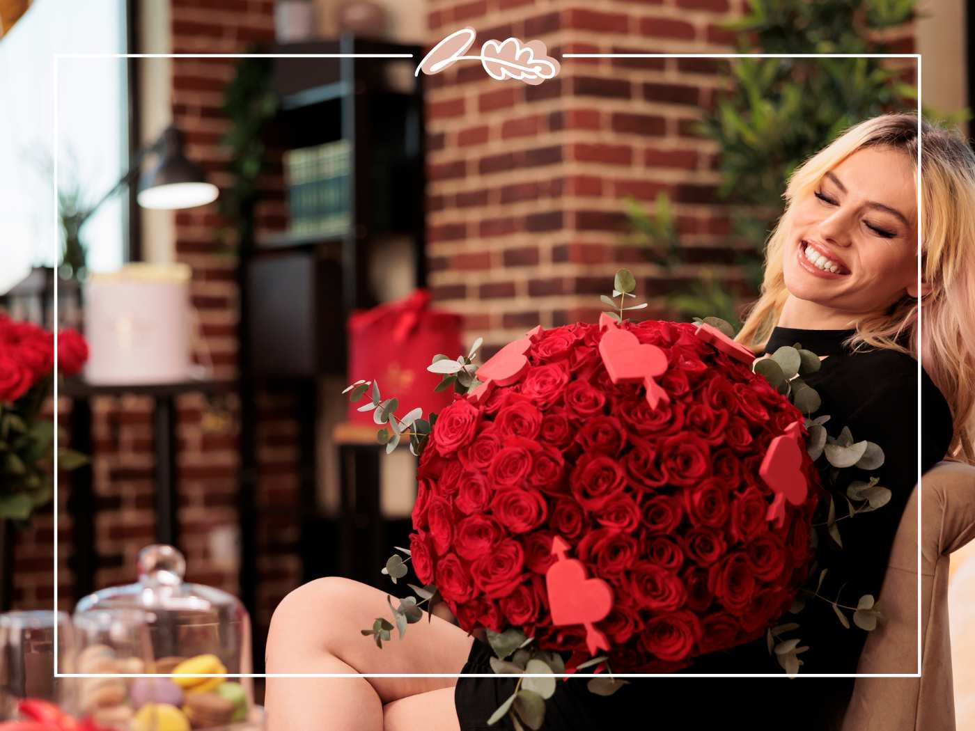 Smiling blonde holds a giant round red rose arrangement with eucalyptus and heart picks in a warm indoor scene.