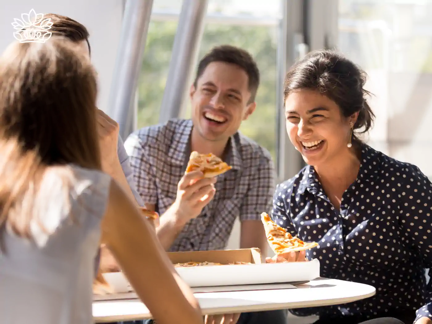Three joyful colleagues enjoying a pizza lunch together in a sunlit office, laughing and sharing a pleasant moment. The ambiance reflects the warmth and camaraderie of a team celebration. Gift boxes for colleagues, delivered with heart by Fabulous Flowers and Gifts.