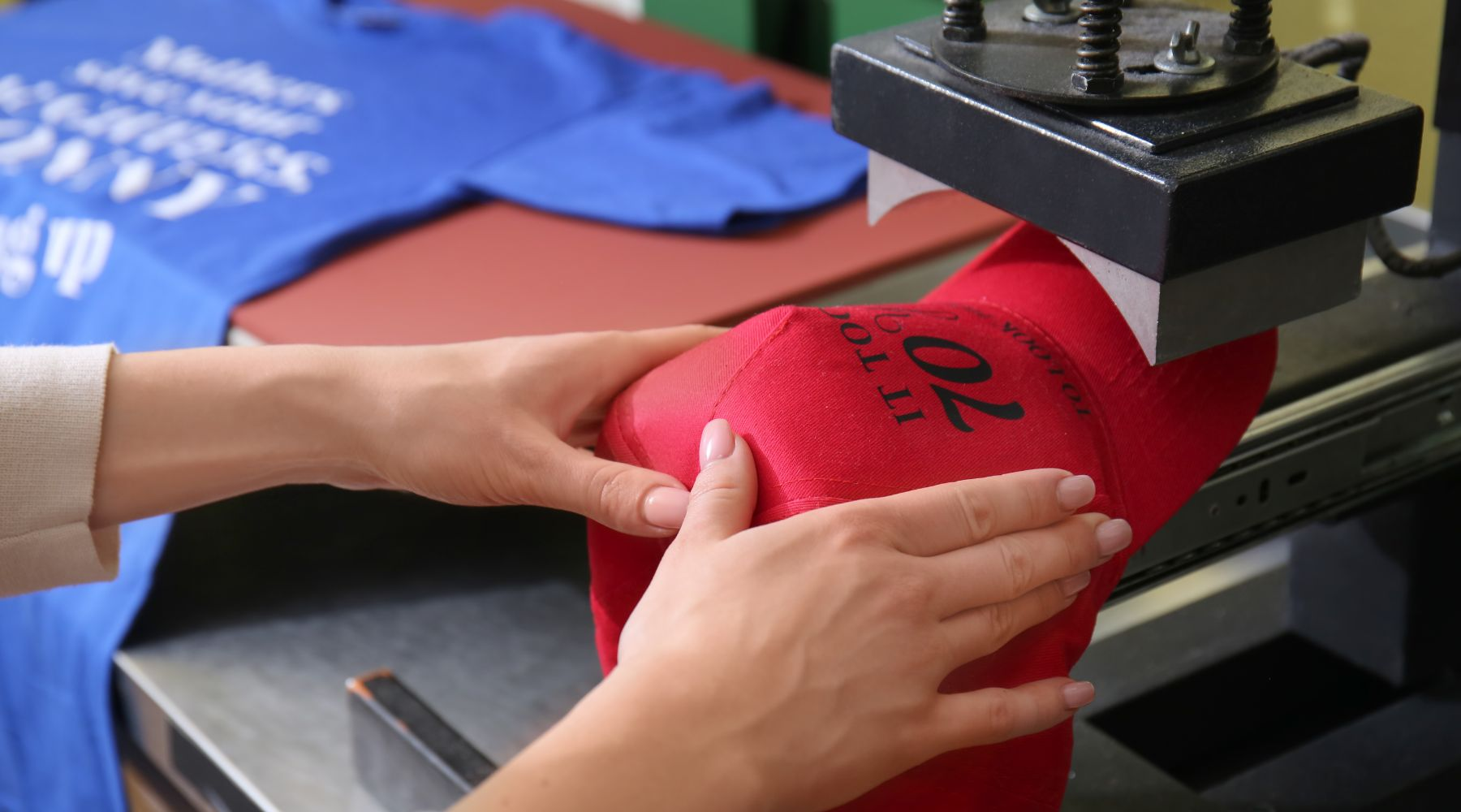 Employee applying a heat press to a custom red baseball cap at a merchandise printing workshop.