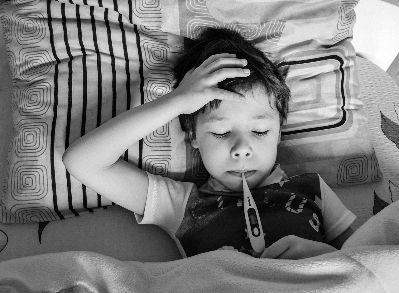 An image of a sick child lying in bed with a thermometer in his mouth.