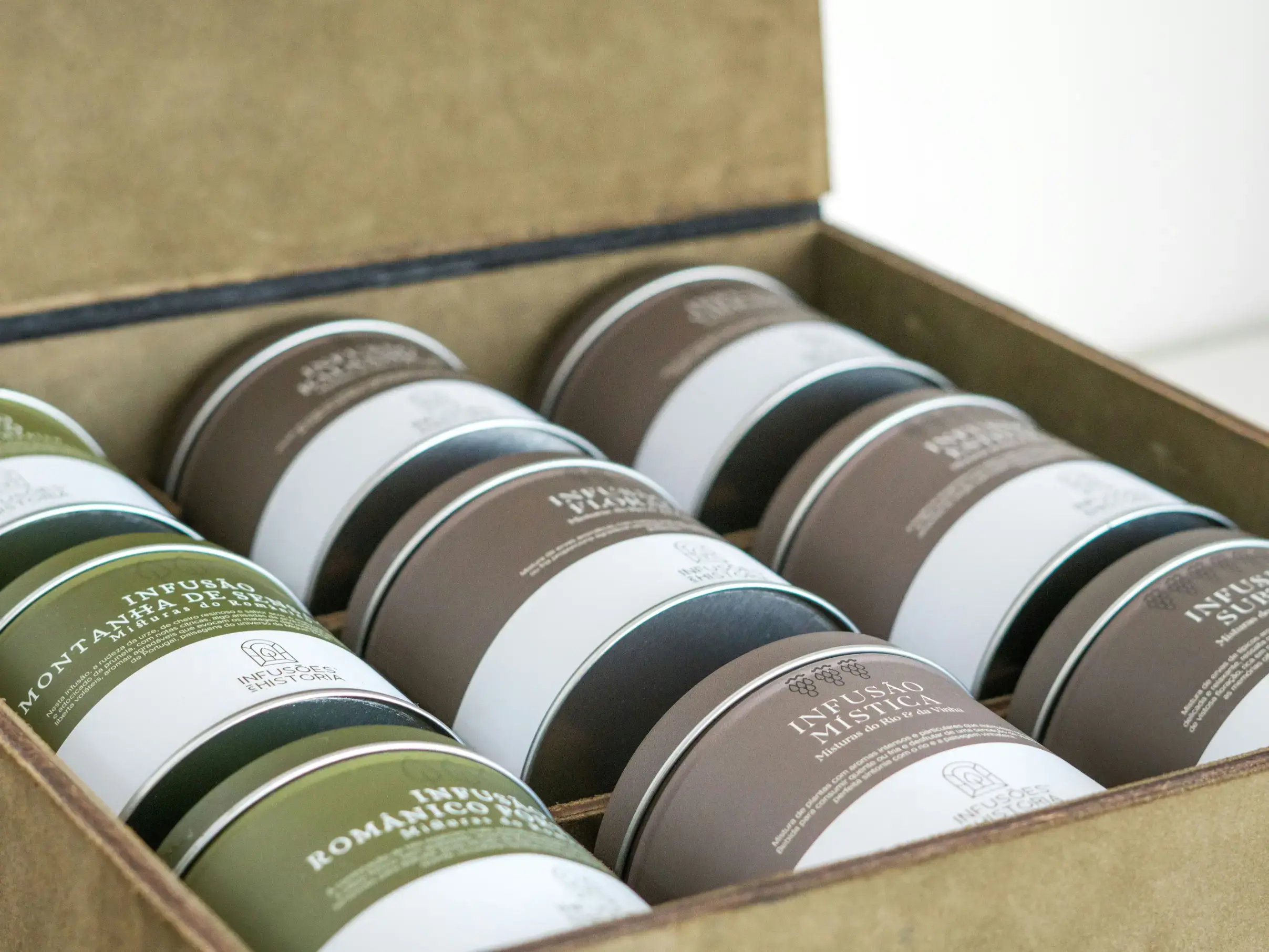 A wooden presentation box is filled with rows of cylindrical tea tins, each featuring minimalist labels with Portuguese text for various herbal infusions. The collection showcases a variety of earth-toned packaging, including shades of forest green and deep brown, arranged neatly to display the brand's artisanal aesthetic.
