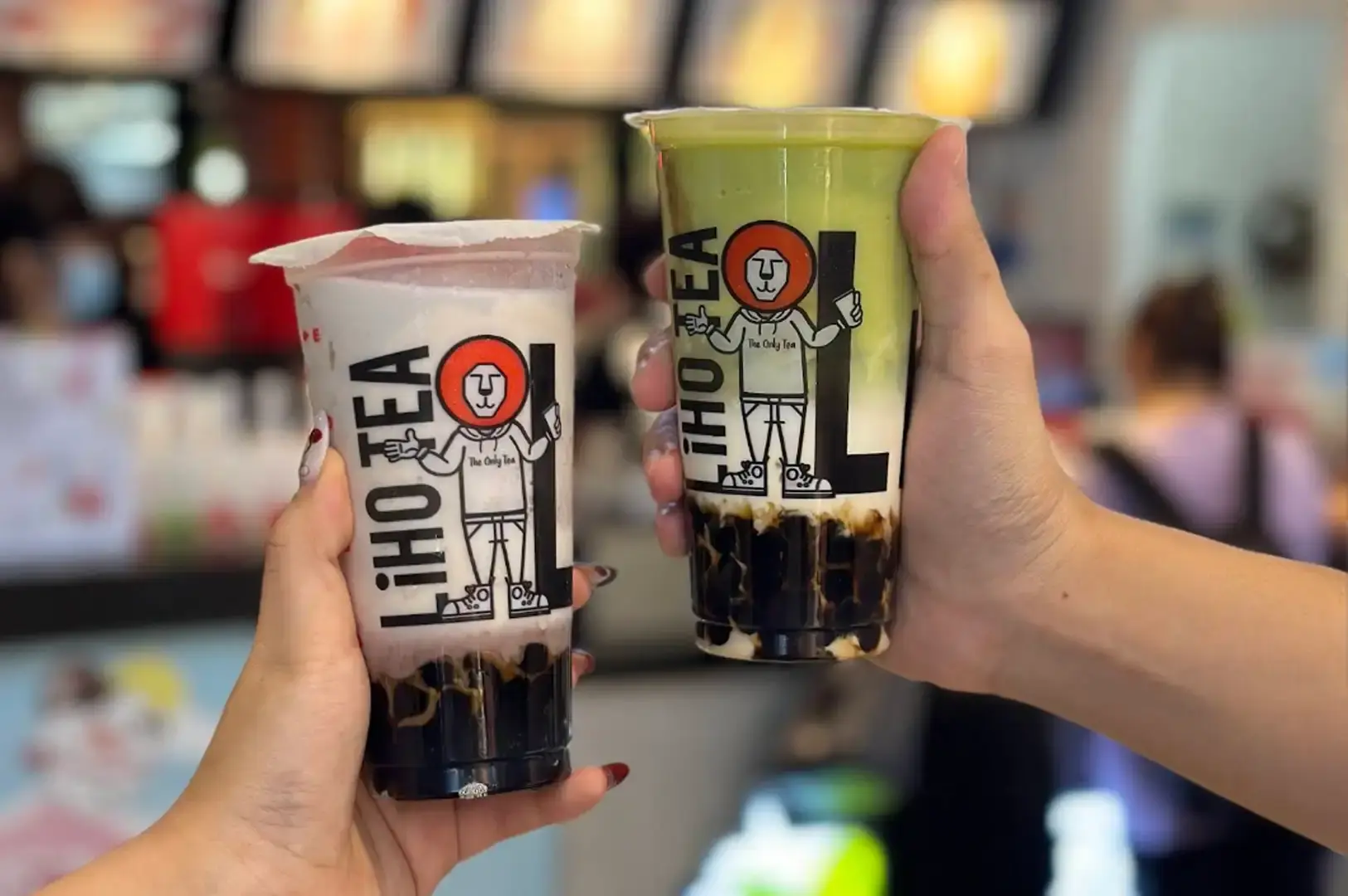 Two hands hold up clear LiHO Tea plastic cups featuring the brand's lion mascot artwork, with the left cup showcasing a pastel purple taro milk tea over dark brown sugar pearls and the right cup presenting a layered matcha green tea over milk and tapioca pearls.