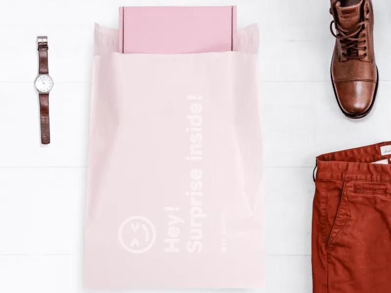 A pastel pink mailing bag with the words "Hey! Surprise inside!" printed in white. A pink box is partially visible, sticking out from the top of the bag. The mailing bag is placed on a clean, white surface. Surrounding the bag are neatly arranged fashion accessories and clothing items: a brown leather wristwatch on the left, a pair of polished brown leather dress shoes at the top right, and a pair of rust-colored trousers at the bottom right. The setup has a minimalist and stylish aesthetic, suggesting a premium or curated unboxing experience. The bag appears to be made of a lightweight, flexible material, used for shipping fashion or lifestyle products.