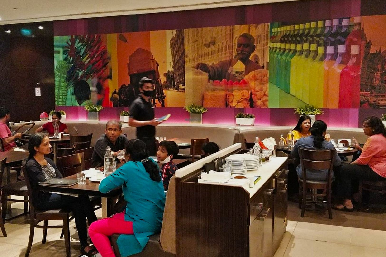 People dine in a casual restaurant with vibrant wall art depicting street scenes. A waiter, masked, serves a family, creating a warm, social atmosphere.