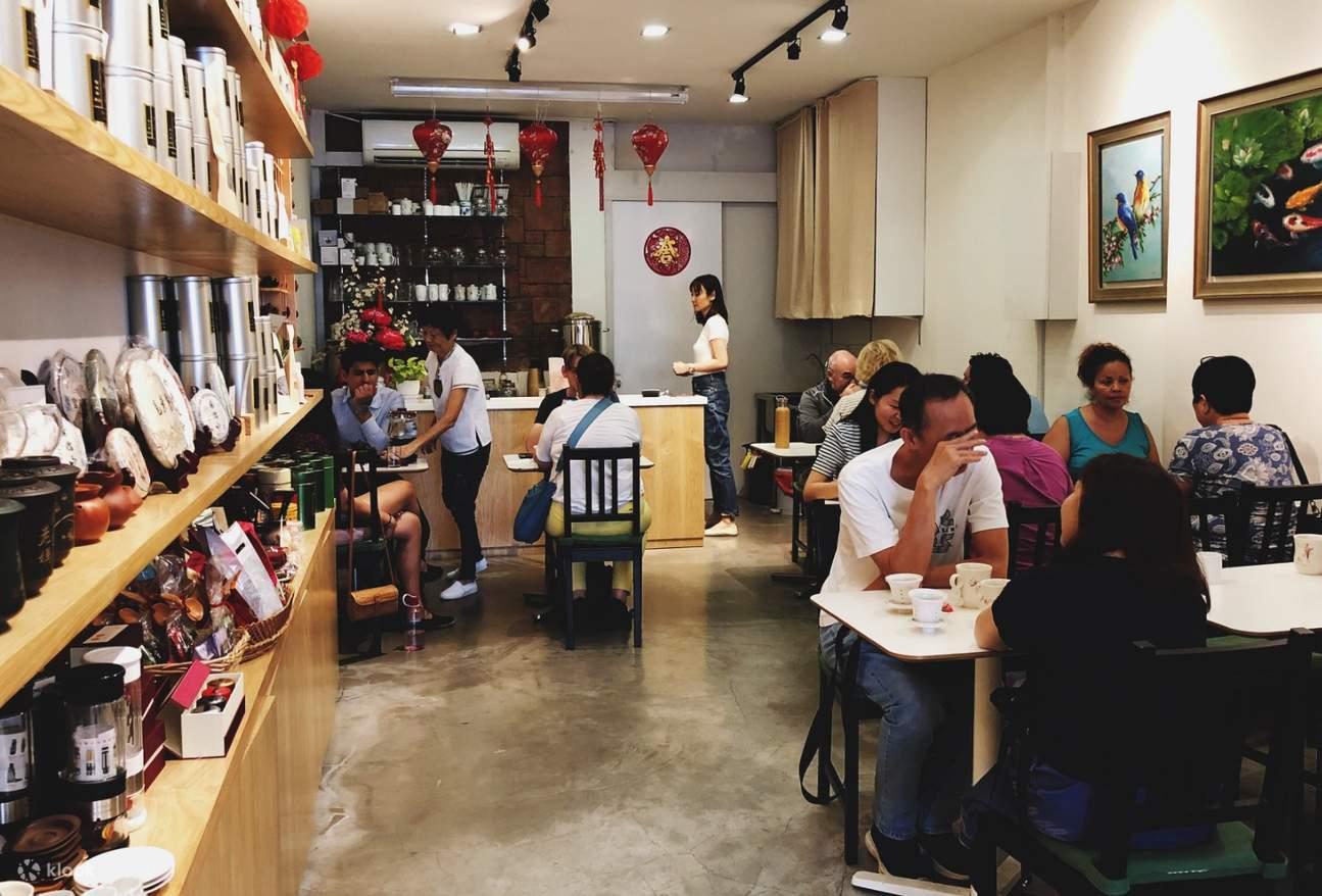 Patrons sit at small tables inside a bustling teahouse featuring shelves lined with canisters and tea cakes. Red lanterns hang from the ceiling, adding a traditional touch to the cozy, narrow interior.