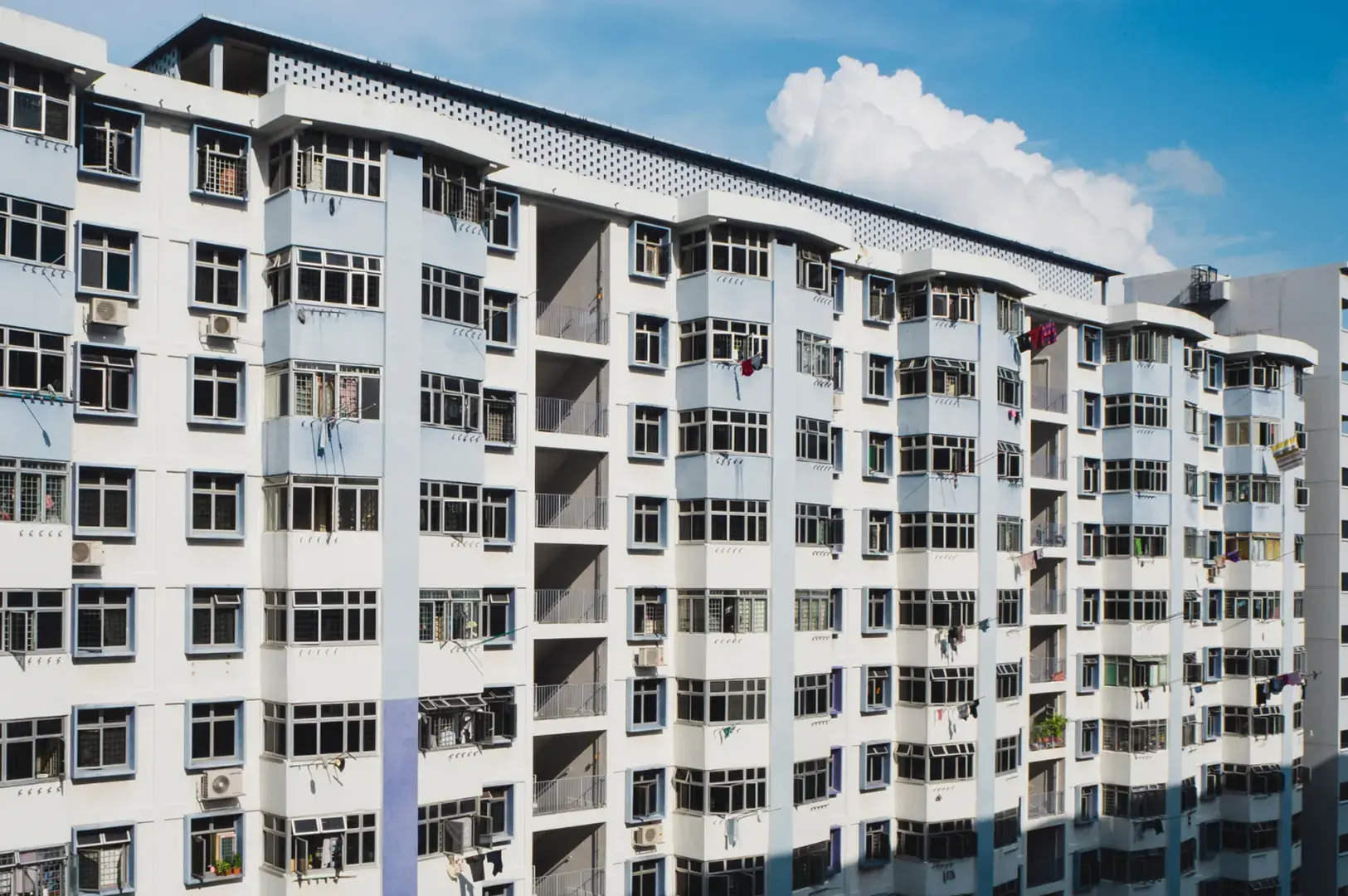A multi-story apartment building with white and light blue facades, many windows, and balconies under a cloudy blue sky.