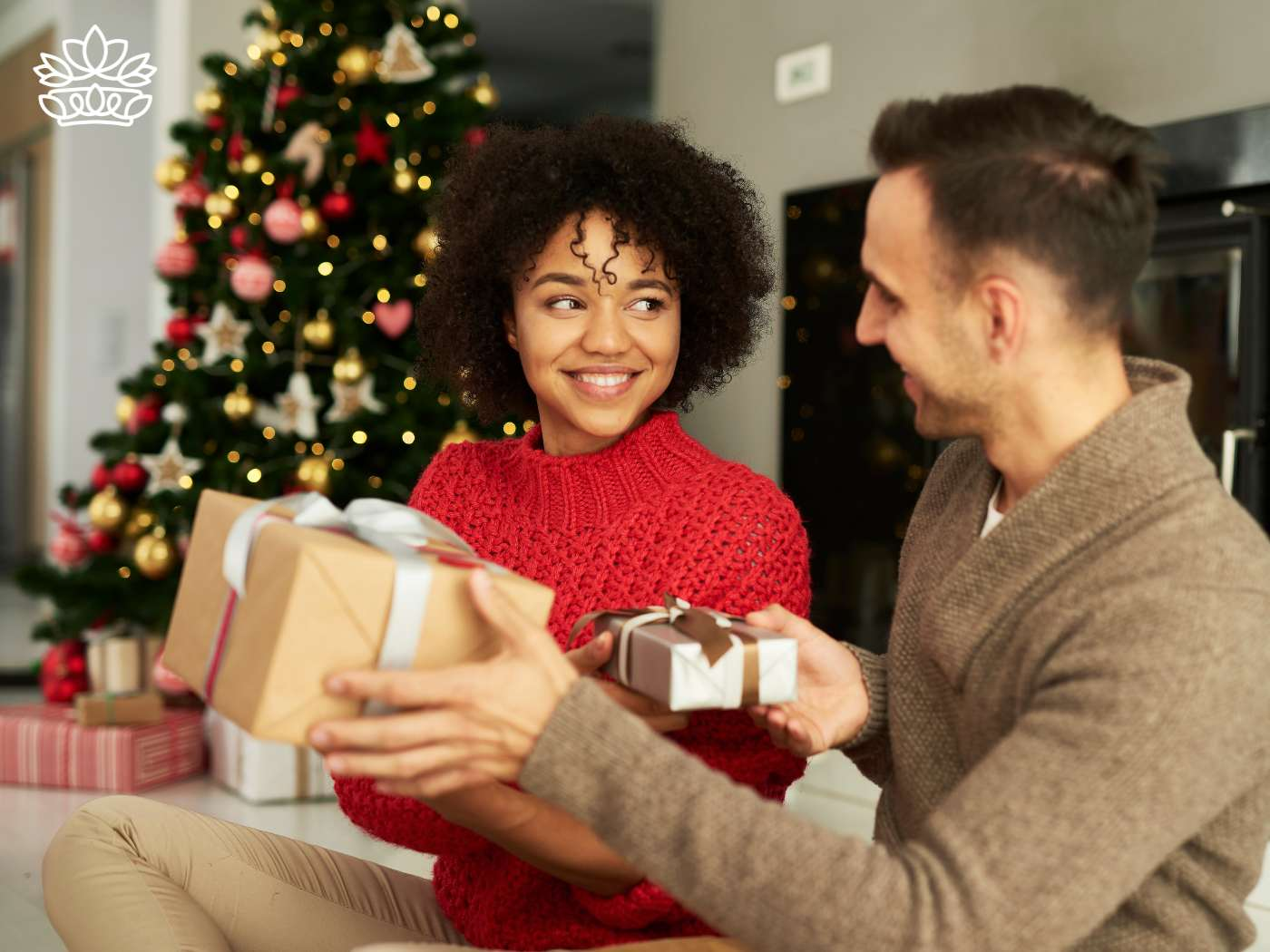 A man surprises a woman with a beautifully wrapped gift hamper, capturing a joyful moment of holiday gift delivery in a cozy setting with a Christmas tree in the background. Part of the Gift Boxes Collection from Fabulous Flowers and Gifts.
