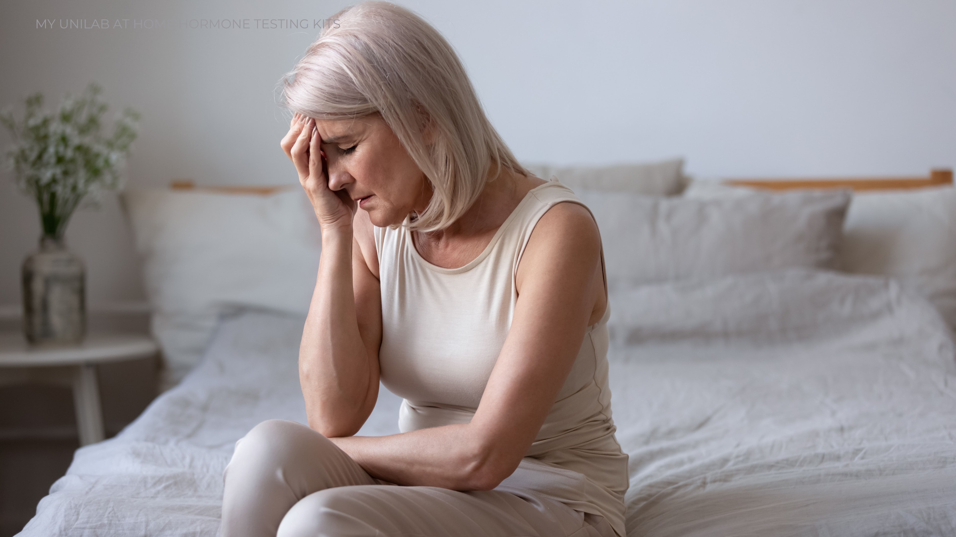 A women going through menopause symptoms sitting at the edge of her bed. Her hand is pressing into her forehead and her facial expression looks weary and in pain.