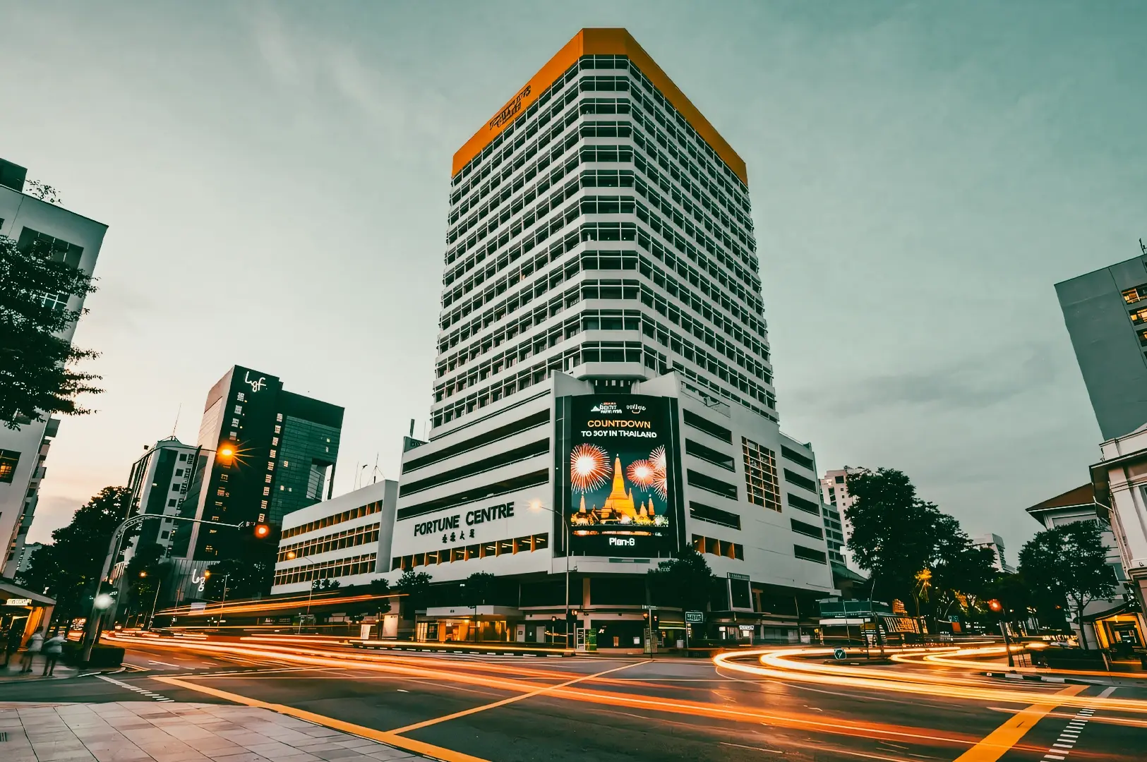 “Wide-angle, low-angle street-level shot of a tall multi-story commercial building at dusk, featuring a white facade with a grid of windows and a contrasting orange rooftop band, surrounded by neighboring urban buildings, with a busy intersection in the foreground captured using long-exposure light trails from passing vehicles, soft ambient evening lighting, and a muted cloudy sky creating a cinematic cityscape atmosphere.