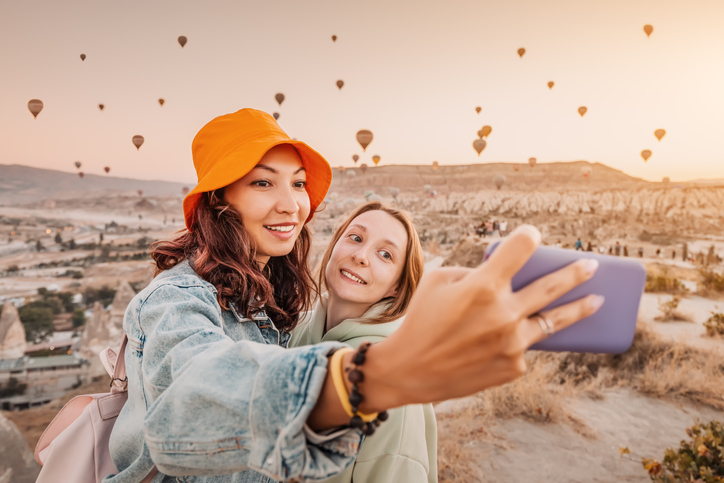 Two happy young women smiling for a selfie with dozens of hot air balloons overhead.