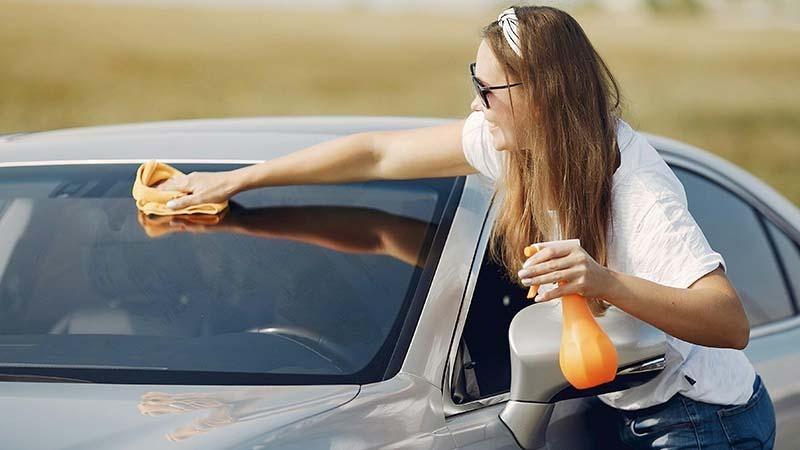 A woman is cleaning her car windshield with an orange microfiber