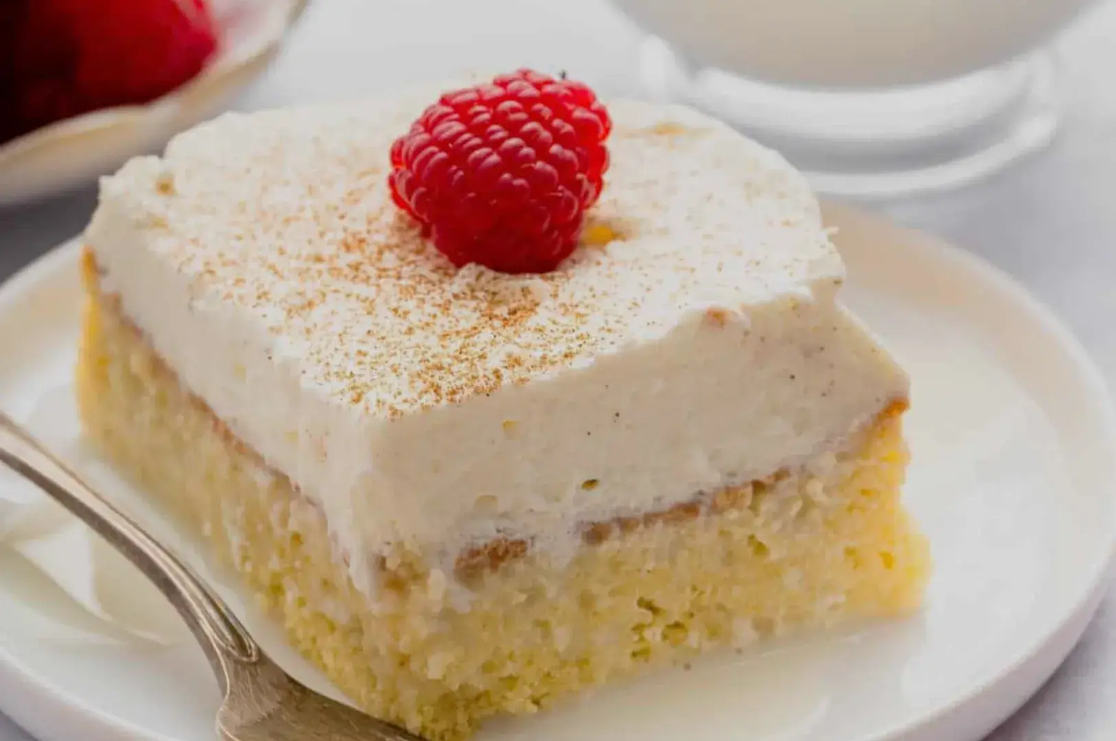 A slice of creamy dessert topped with whipped cream and a raspberry, served on a white plate with a spoon.