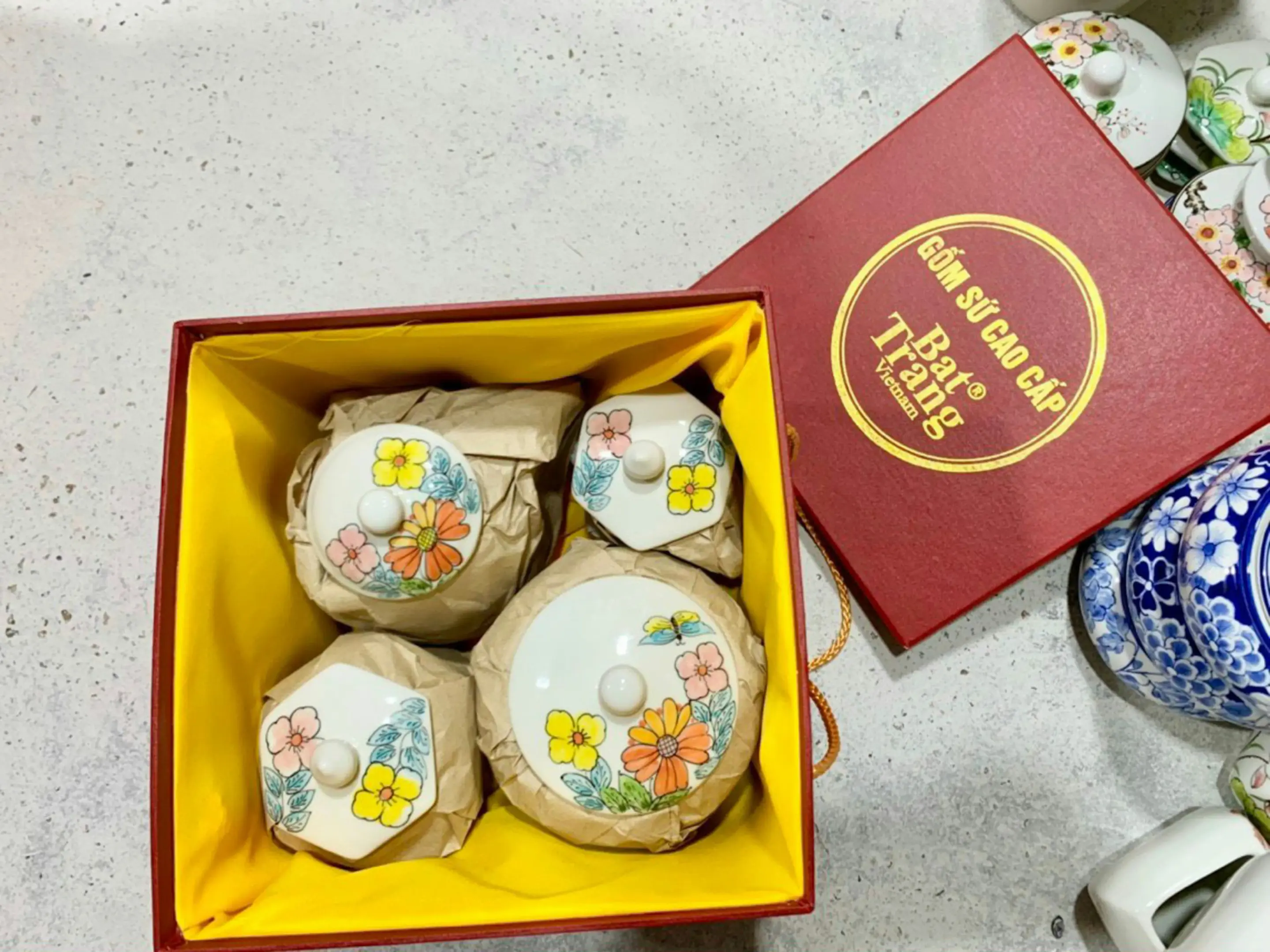 This image displays a set of delicate Bat Trang Vietnamese ceramic jars decorated with colorful floral patterns and nestled in a yellow-lined gift box. The red lid of the box sits nearby, featuring elegant gold lettering that highlights the high-quality craftsmanship of the pottery.