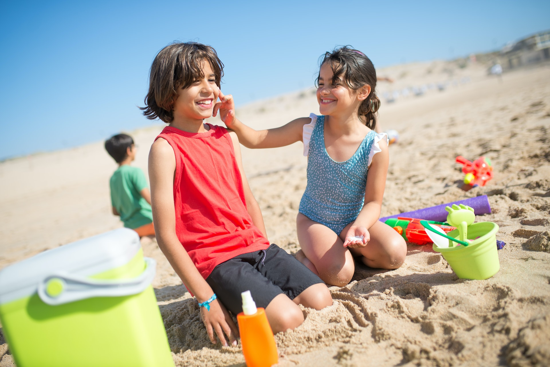 Casal de crianças sentado na praia com brinquedos para mexer na areia. A menina sorri enquanto passa protetor solar no rosto do menino. Ambos parecem ter entre 7 a 9 anos de idade, tem pele clara e cabelos castanhos escuros. Imagem - Pexels.com
