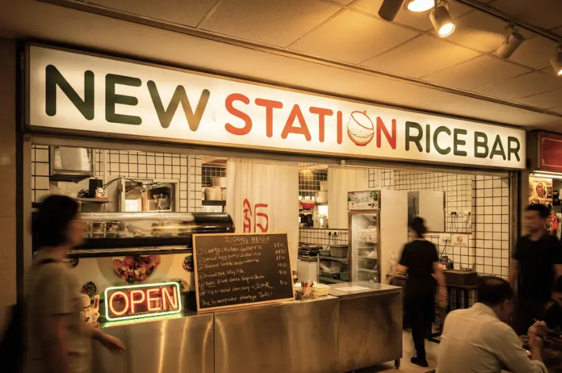 The brightly lit storefront of New Station Rice Bar inside Fortune Centre, featuring a large sign with green and orange lettering, a glowing neon "OPEN" sign, a handwritten daily menu on a chalkboard, and patrons dining at a stainless steel counter while others walk past.