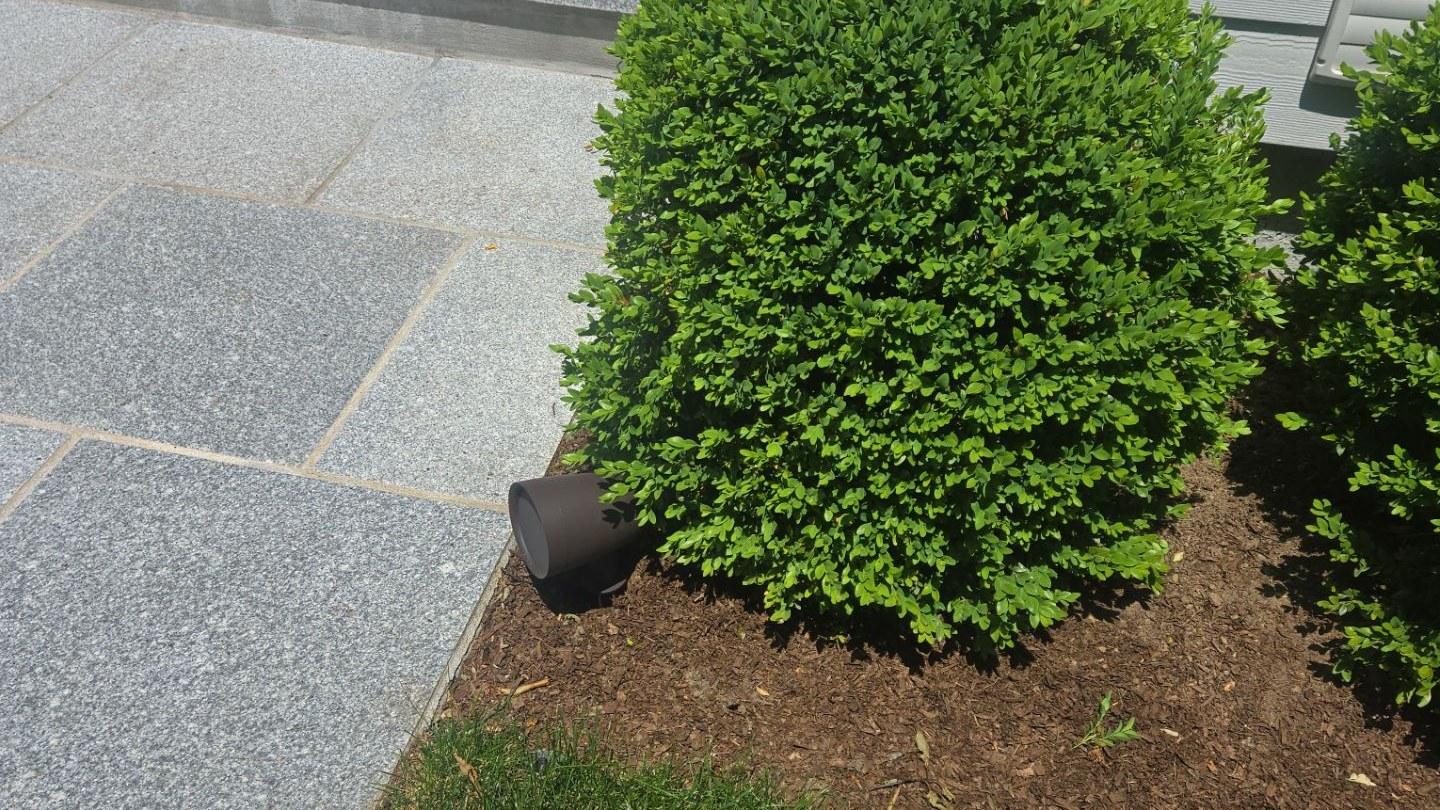 A staked landscape speaker with black finish beside a boxwood where a granite patio meets the home's siding.