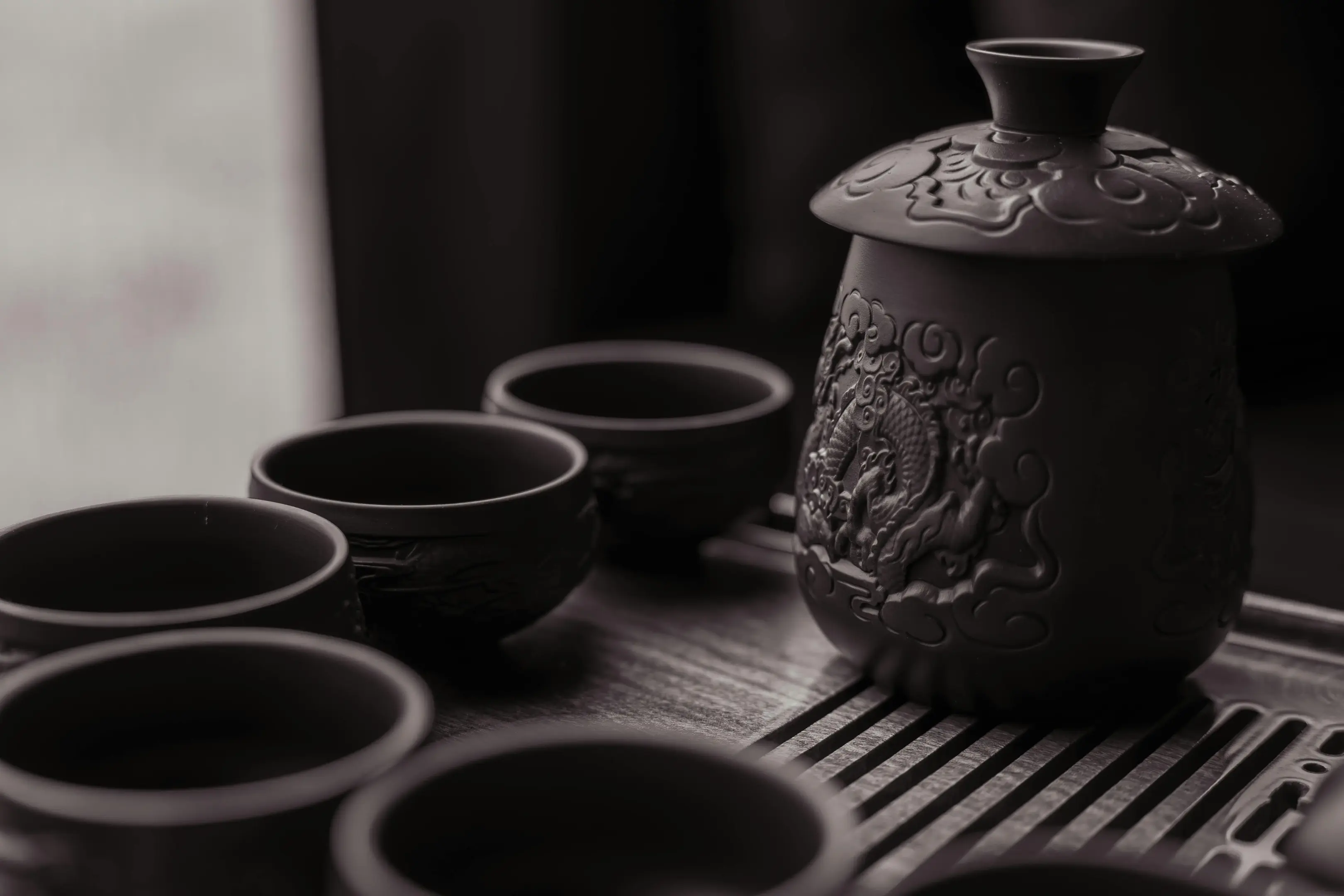 A set of dark, intricately carved ceramic teaware sits atop a slatted wooden tray. The collection features a tall lidded tea vessel adorned with a dragon motif alongside several matching small cups.