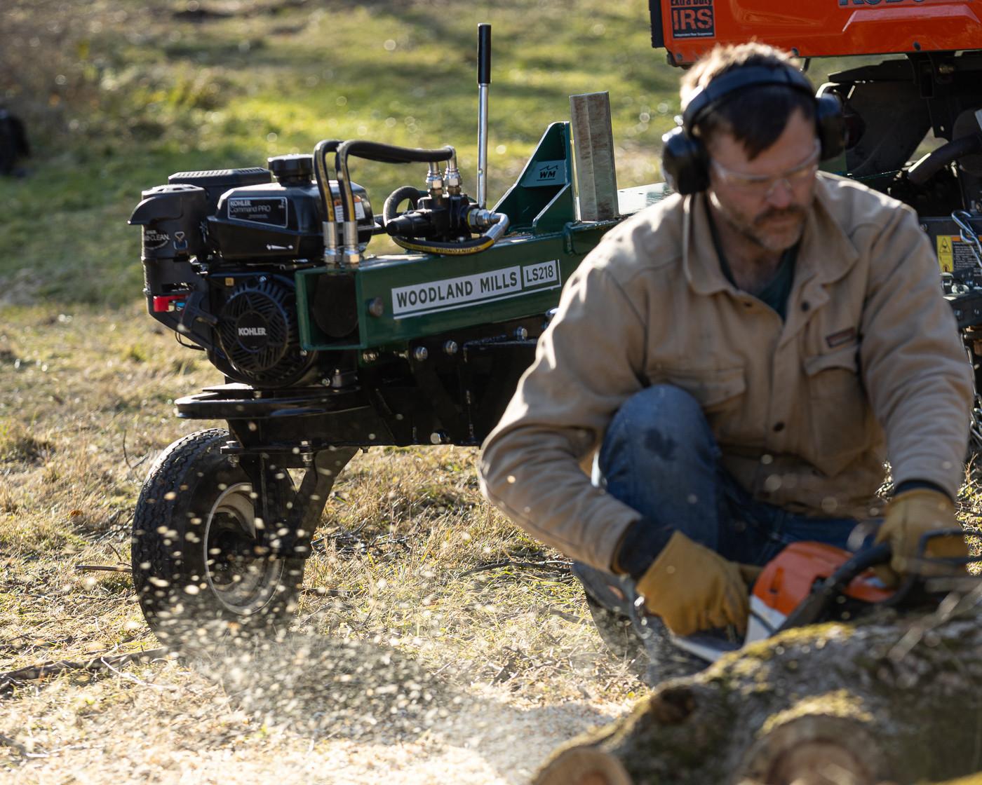 A man cutting wood with a chainsaw in front of a Woodland Mills LS218 2-Way Log Splitter.