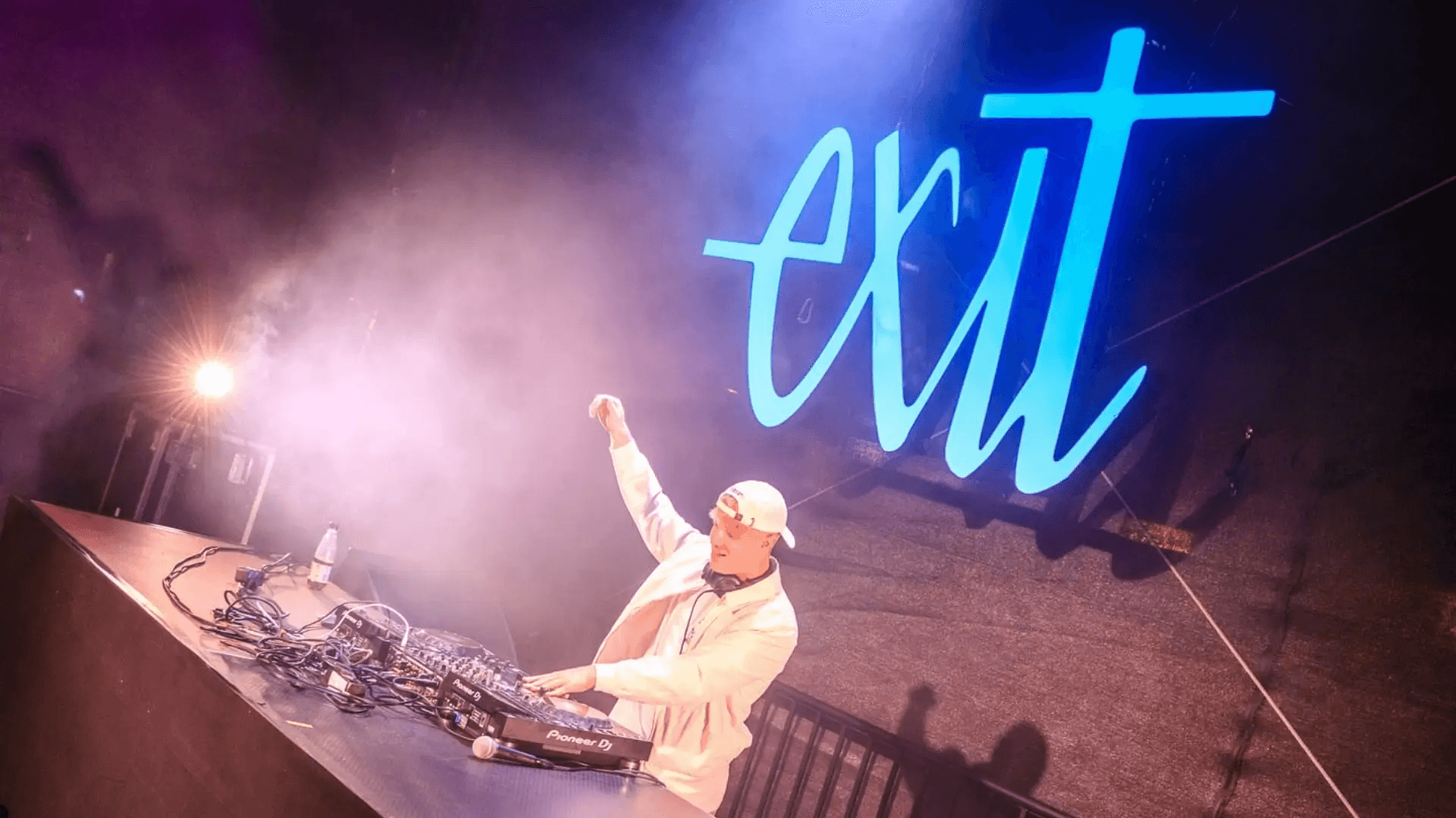 DJ Nathan White steht auf einer beleuchteten Bühne hinter einem Mischpult, hebt eine Hand in die Luft und legt elektronische Musik auf; im Hintergrund leuchtet ein großes blaues Logo, umgeben von Nebel und Bühnenlicht.