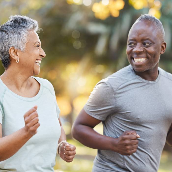 An elderly couple joyfully jogging in a park, the woman in a light green top and the man in grey, both smiling and enjoying their exercise together, representing a healthy lifestyle promoted by The Good Stuff Health Shop South Africa, TERRA NOVA Collection.